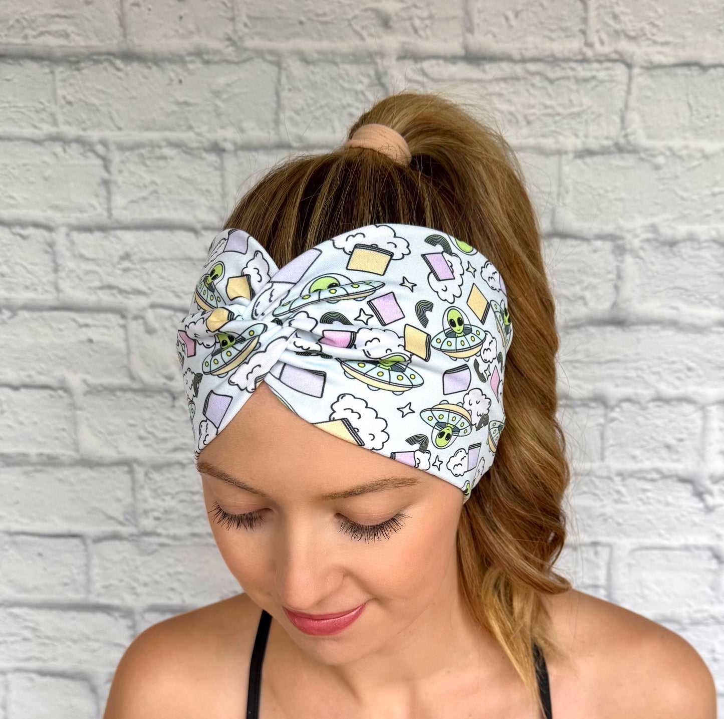 Woman with hair in ponytail wearing wide, twisted, light blue headband with alien theme print.