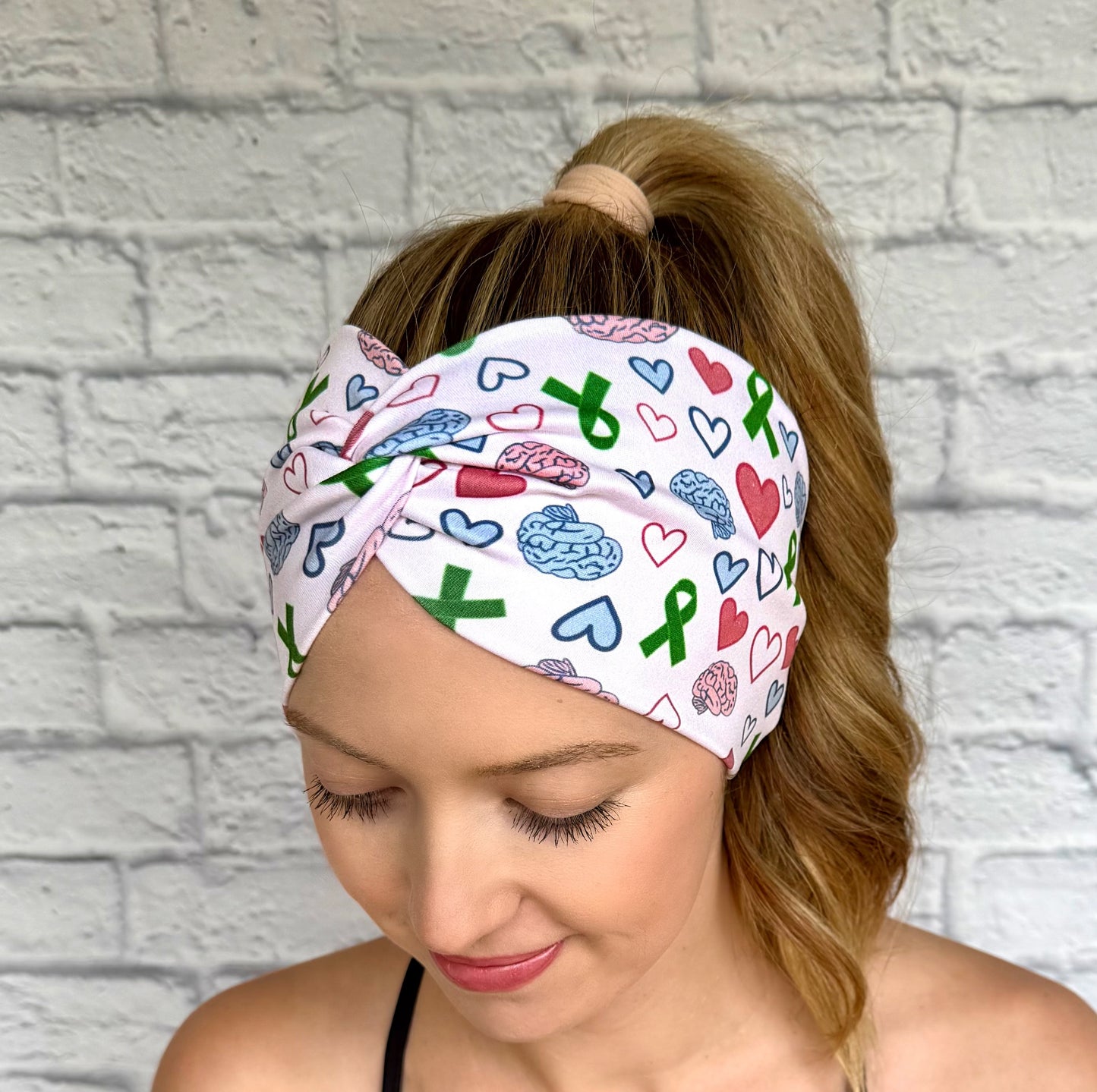 Woman with hair in curly ponytail wearing wide, twisted headband with green ribbon, pink and blue brain, and pink and blue heart print.