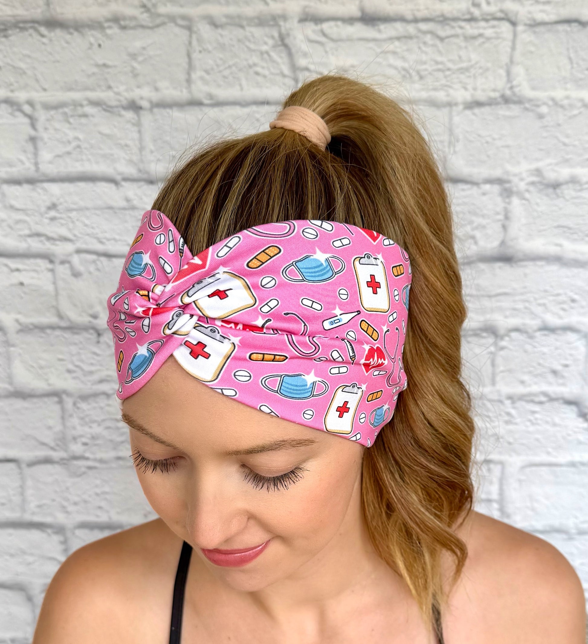 Woman with hair in ponytail wearing wide, twisted, stretchy headband in pink with medical theme print.