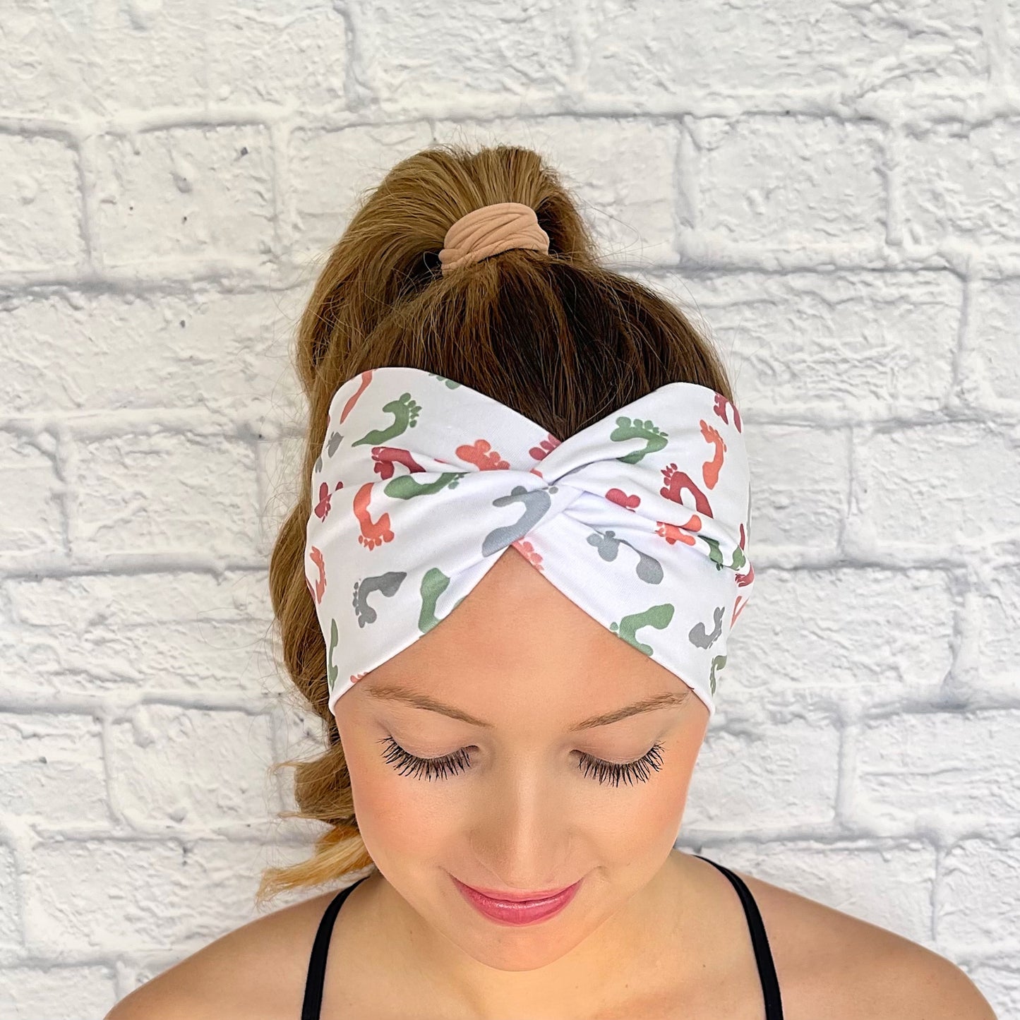 Woman with hair in curled ponytail wearing wide, twisted, white headband with peach, dark pink, teal, and dark blue baby feet print.