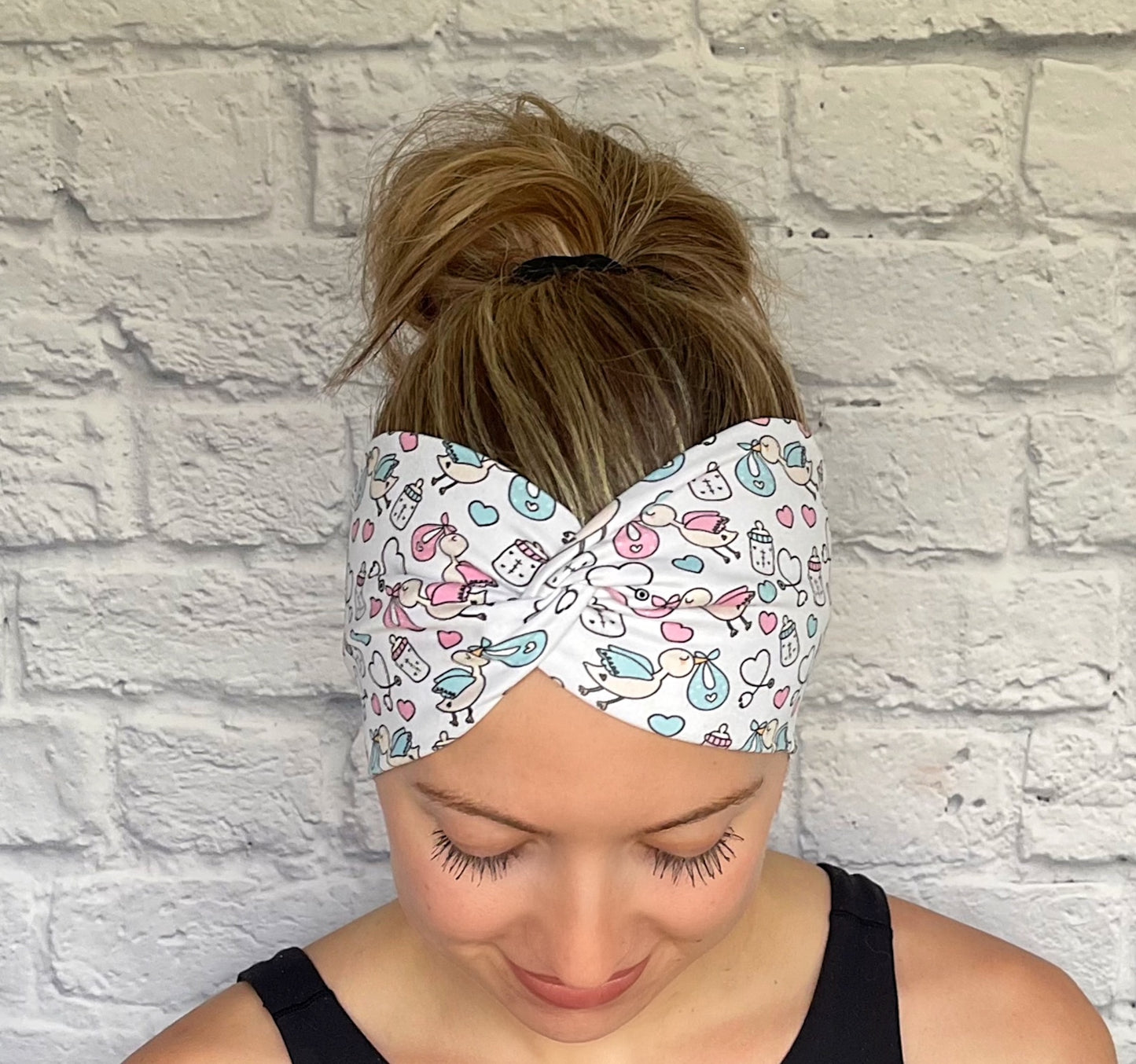 Woman with hair in bun wearing stretchy, wide, twisted headband in white with baby theme print- storks, hearts, bottles, and stethoscopes.