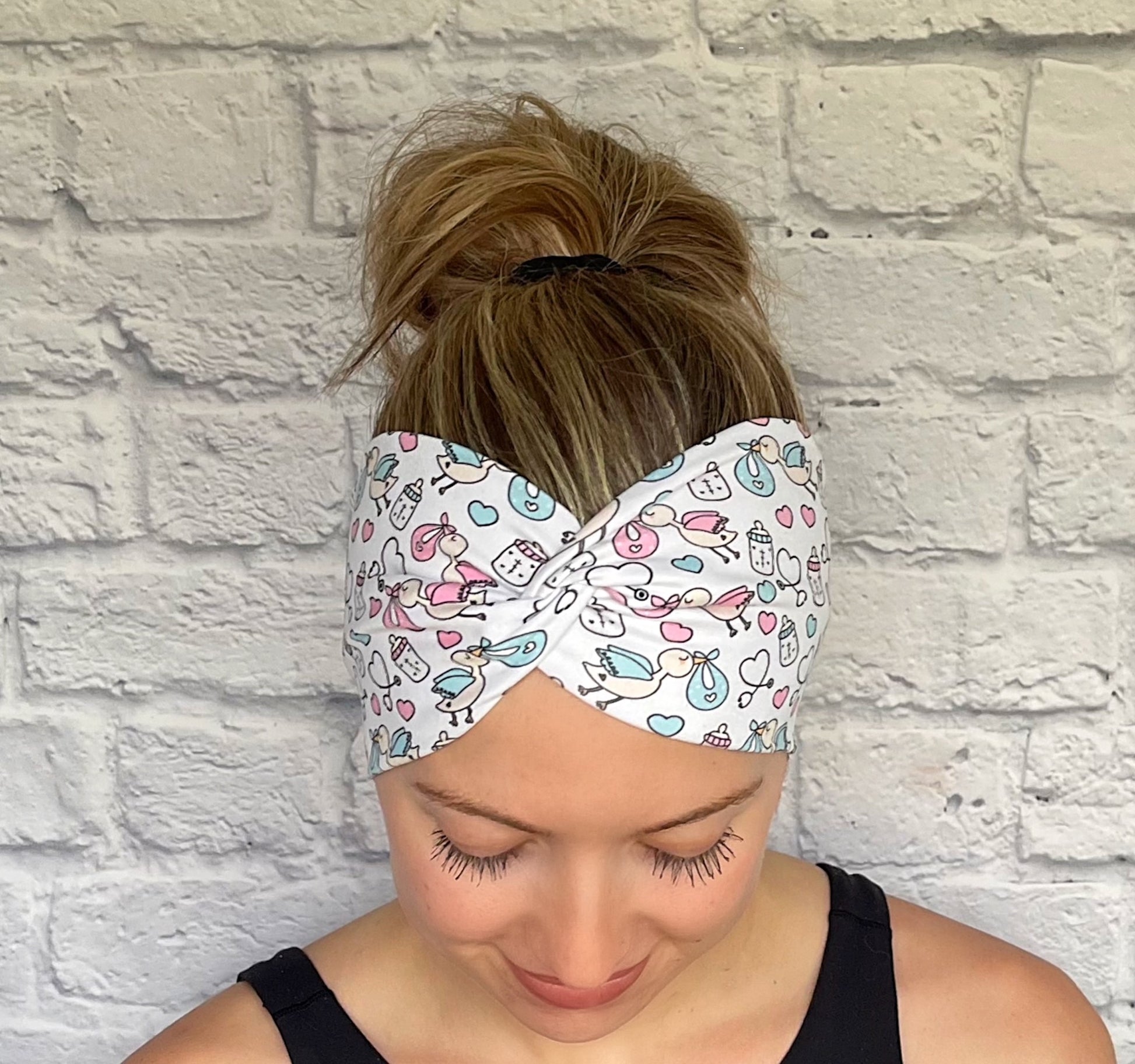 Woman with hair in bun wearing stretchy, wide, twisted headband in white with baby theme print- storks, hearts, bottles, and stethoscopes.