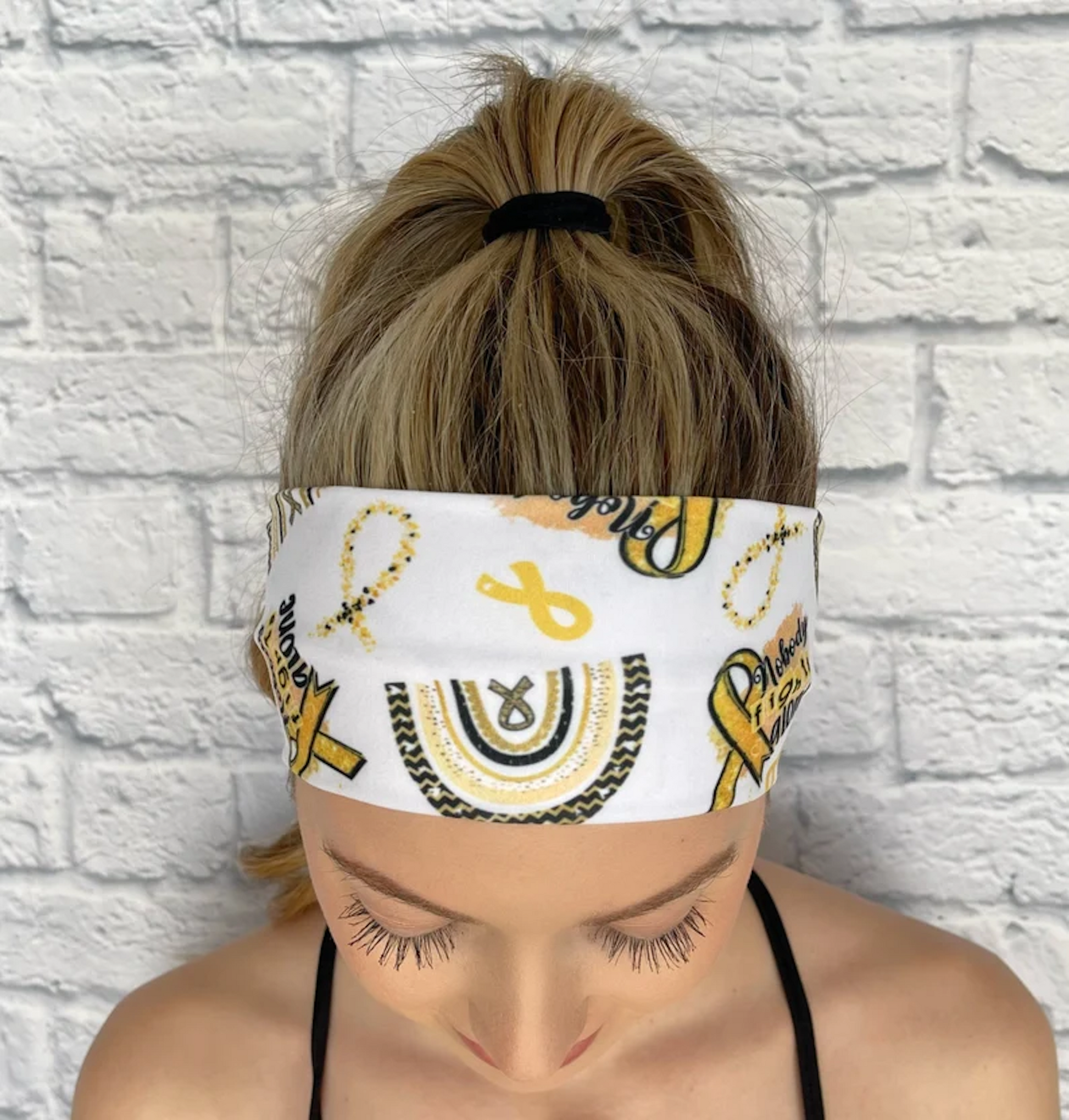 Woman with hair in curled ponytail wearing flat, stretchy headband in white with "no one fights alone" print, yellow/gold rainbow, and yellow/gold ribbon print.