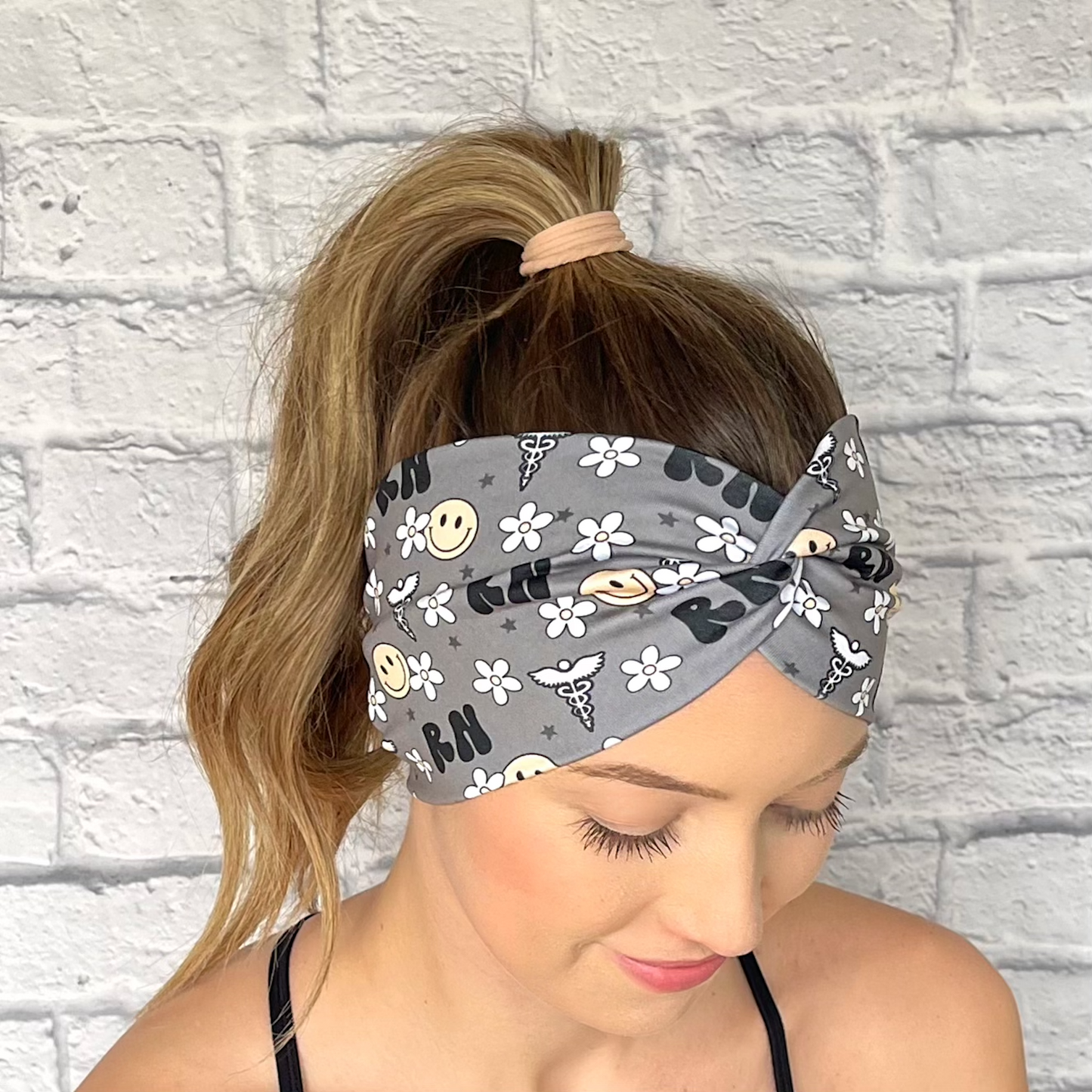 Woman with hair in curled ponytail wearing wide, twisted headband. The headband is gray with "RN" print, flowers, smiley faces, and medical theme print in black, white, and yellow.