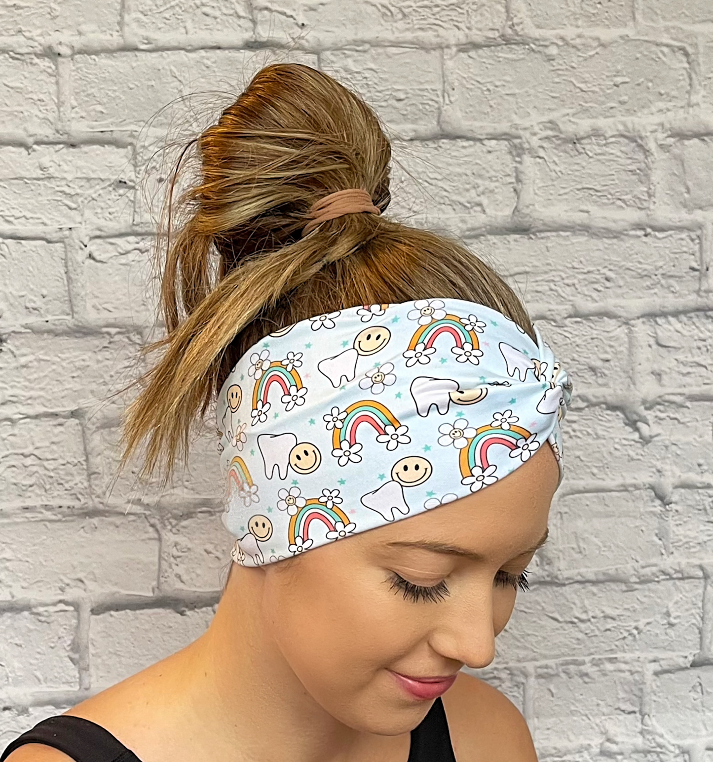 Woman with hair in bun wearing wide, twisted headband in light blue with teeth, rainbows, flowers, and smiley faces print.