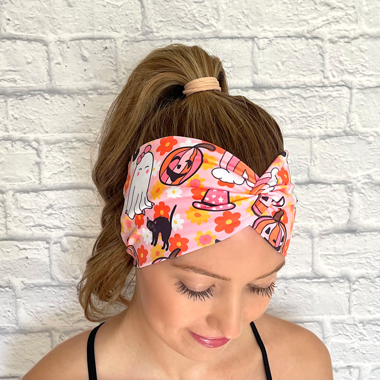 Woman with hair in curled ponytail wearing wide, twisted headband in pink with floral, rainbow, and halloween print in pink, white, black, yellow, and orange.