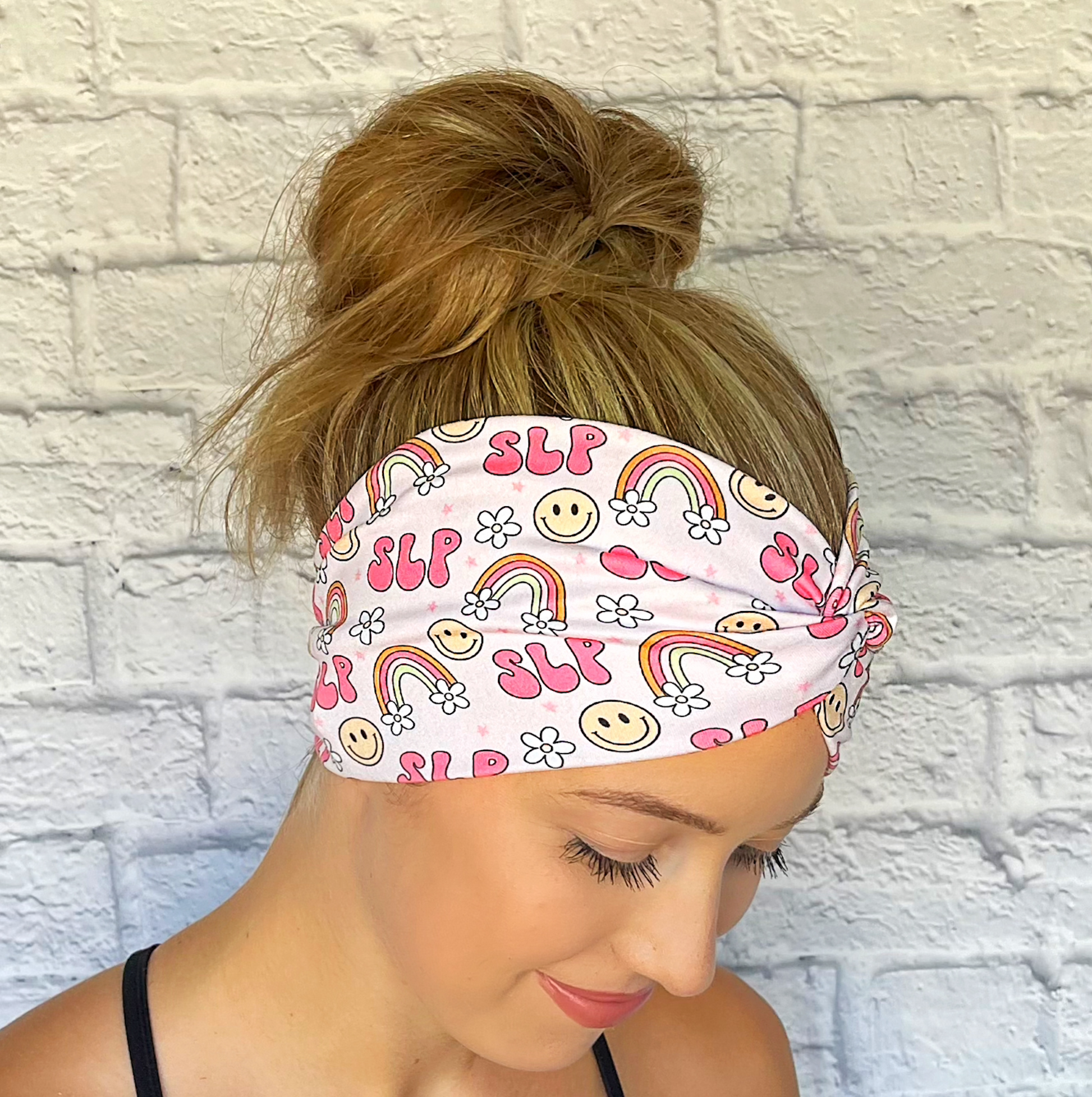 Woman with hair in bun wearing wide, twisted pink headband stating "slp", with rainbows, smiley faces, and white flowers.
