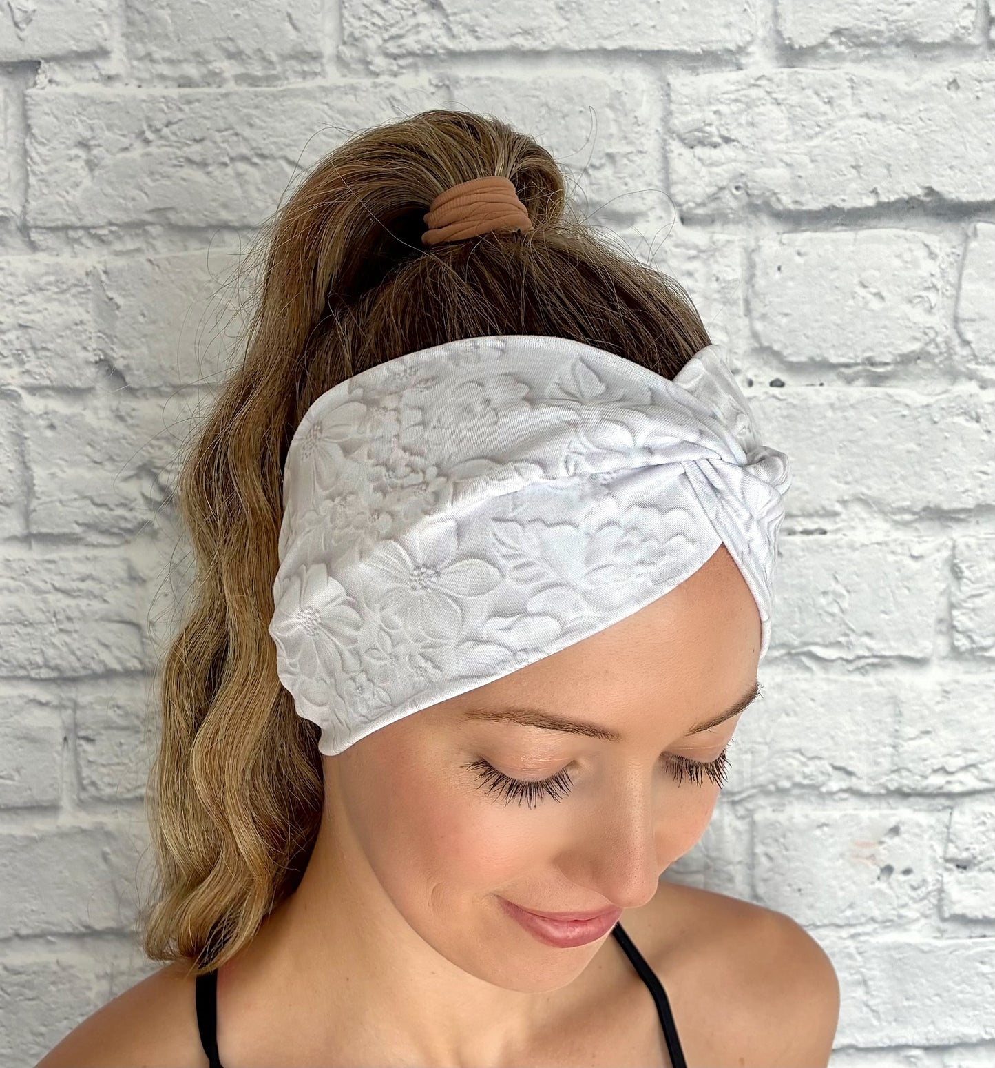 Woman with hair in ponytail wearing wide, twisted, white headband with faux embossed floral print and gray shadowing.