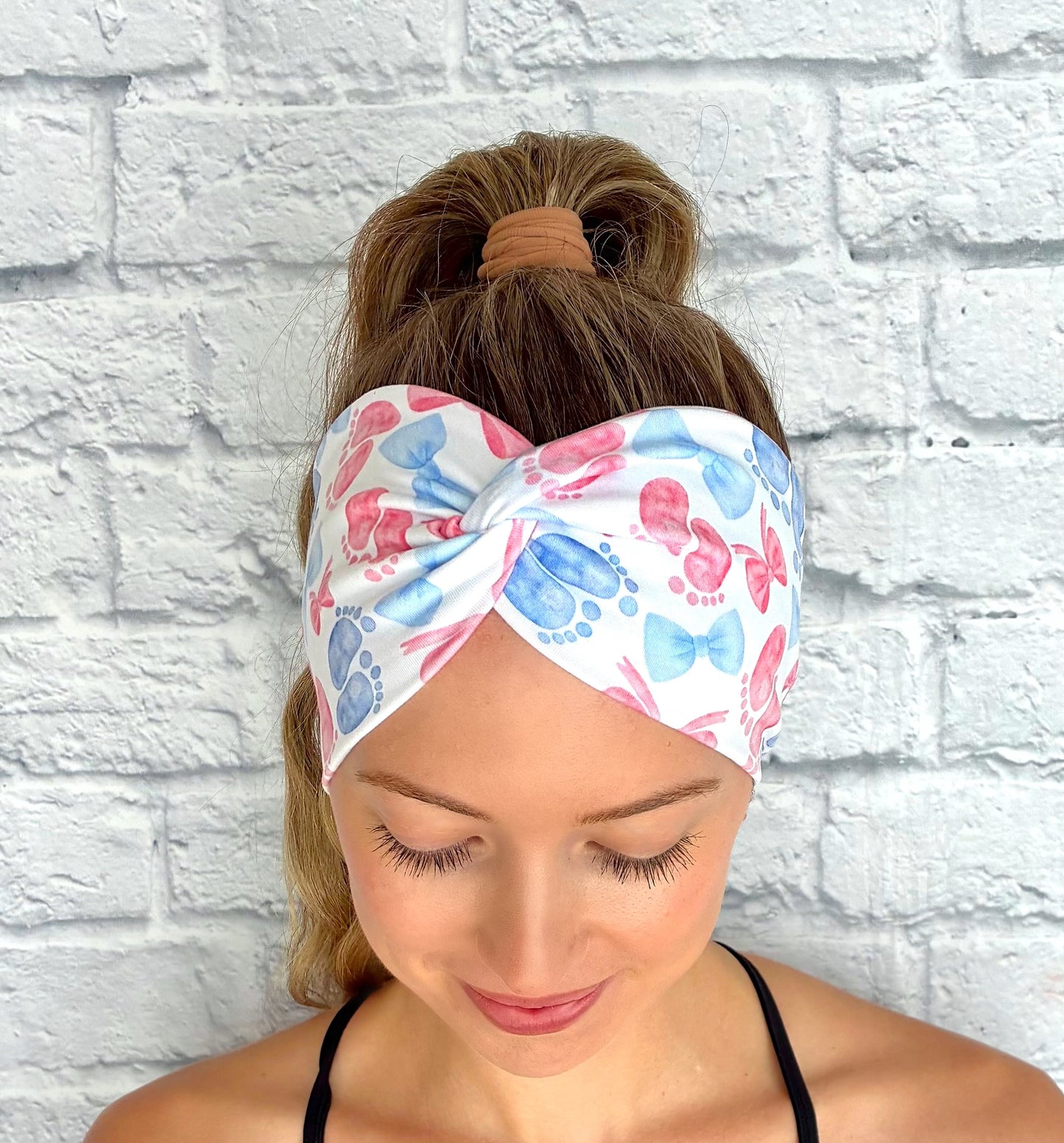 Woman with hair in ponytail wearing wide, twisted, white headband with pink and blue baby feet and bow print.