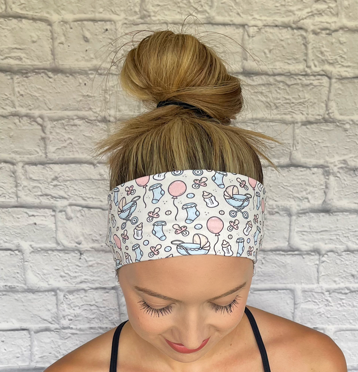 Woman wearing a cream color headband with baby-themed design against a white brick wall.