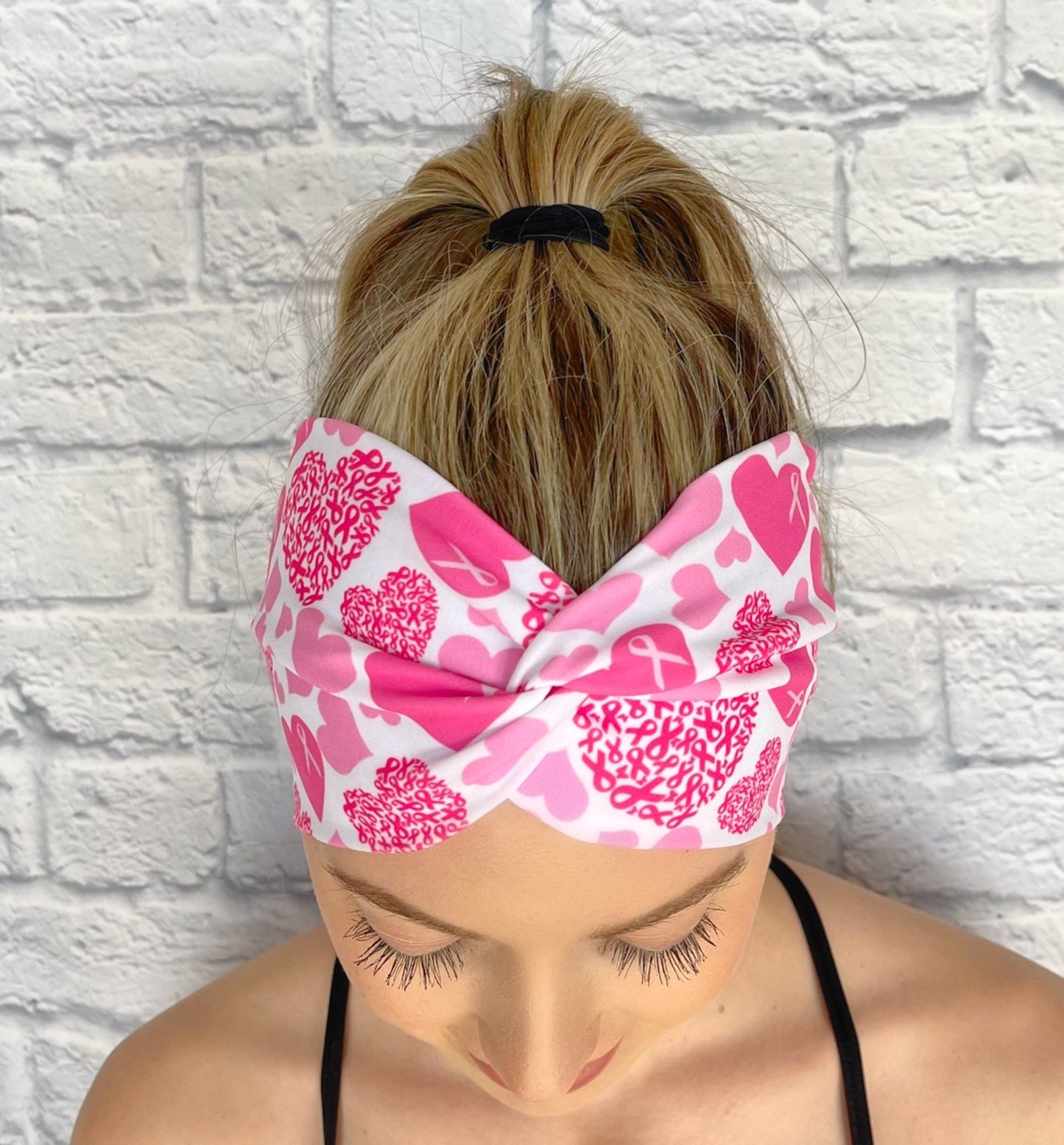 Woman with hair in ponytail wearing wide, twisted headband in white with pink heart and pink ribbon print.