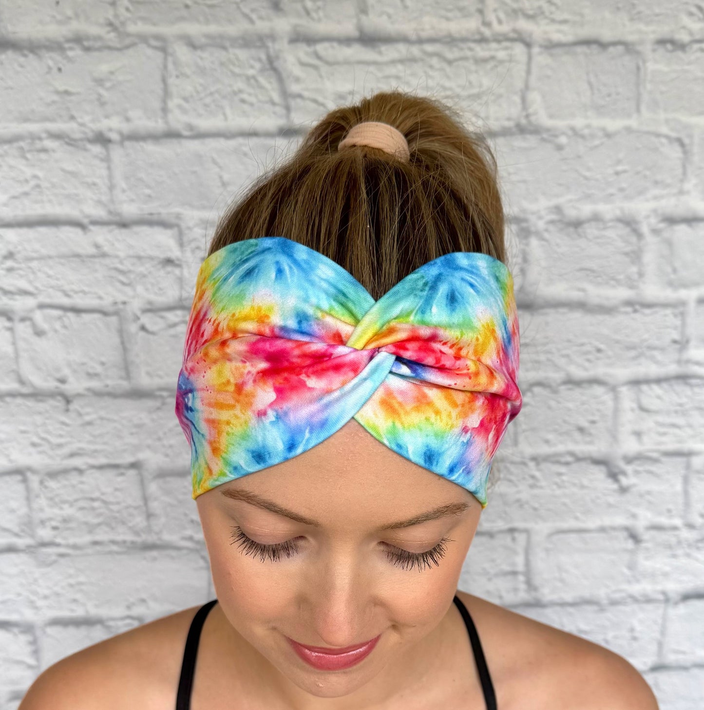 Woman with hair in ponytail wearing wide, twisted headband with rainbow color tie dye print.