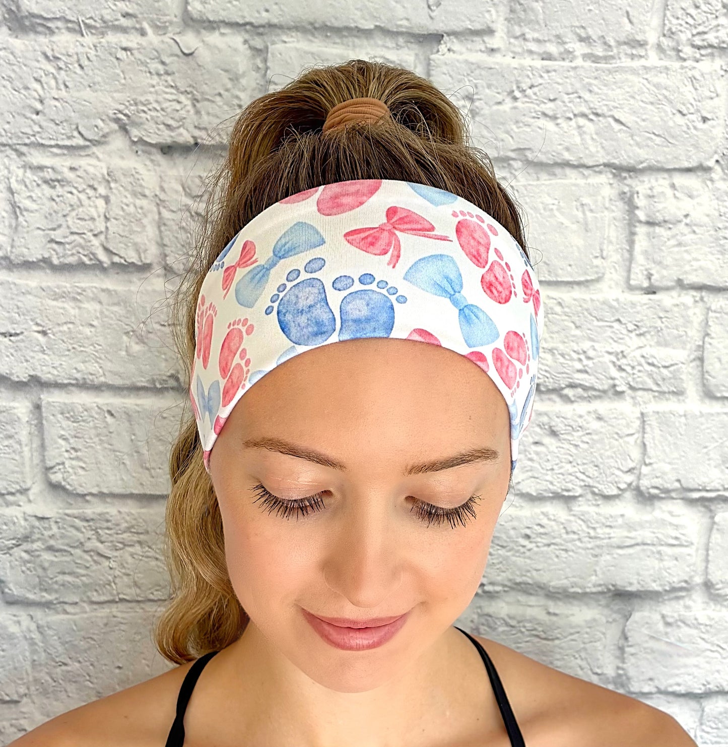 Woman with hair in ponytail wearing flat, stretchy, white headband with pink and blue baby feet and bow print.