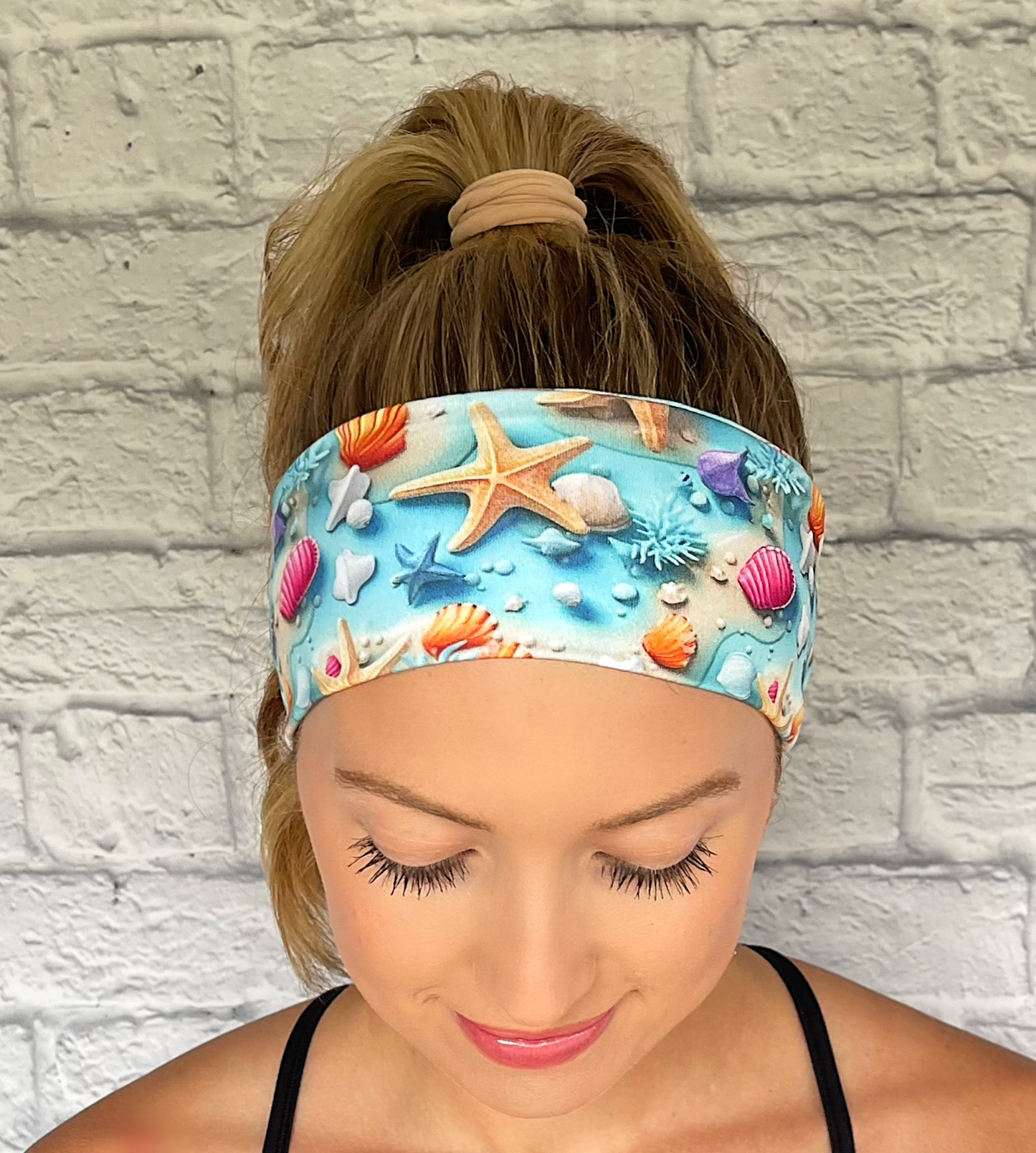 Woman with hair in ponytail wearing light blue headband with ocean theme print- water, sand, starfish, and shells.