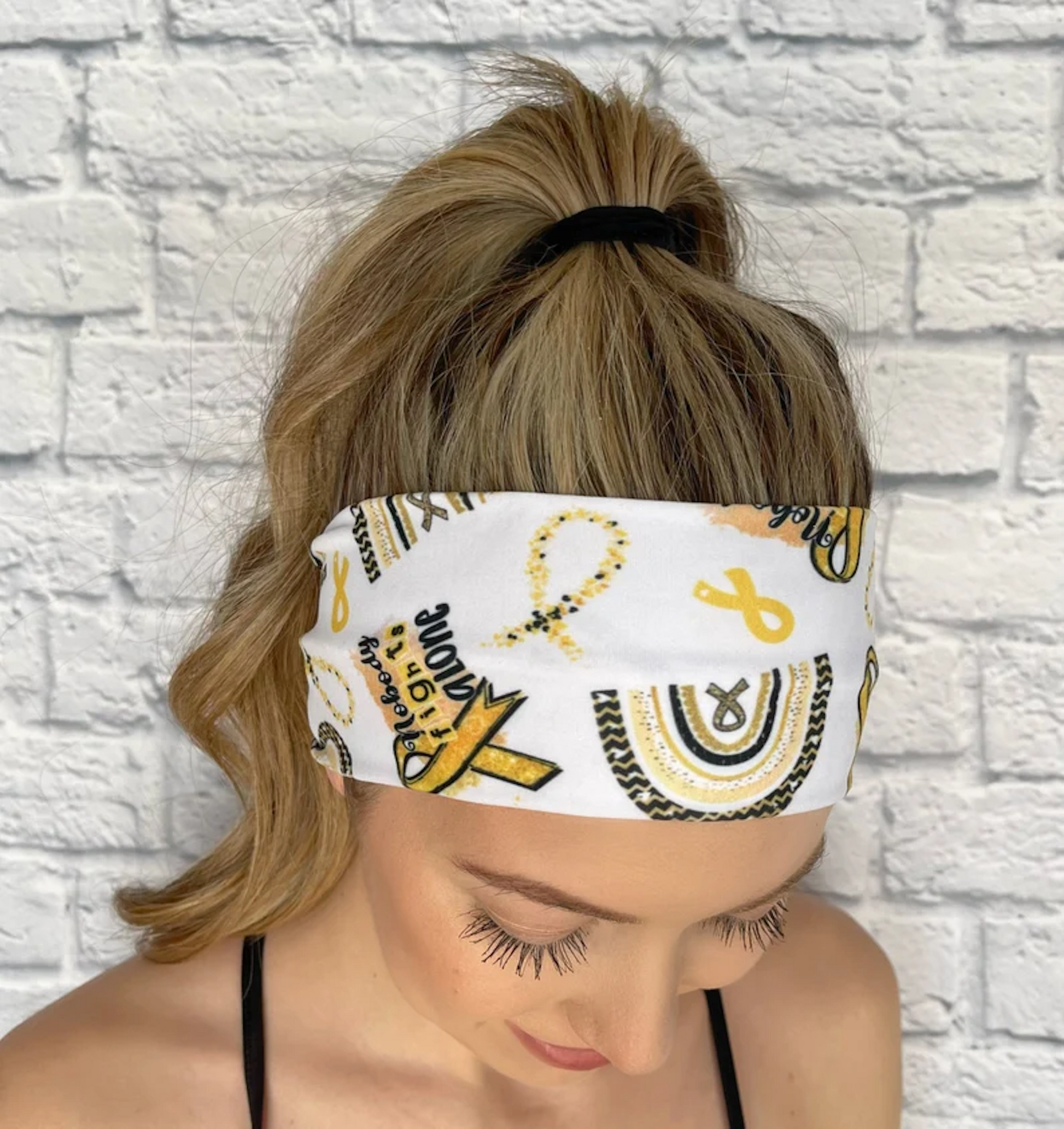 Woman with hair in curled ponytail wearing flat, stretchy headband in white with "no one fights alone" print, yellow/gold rainbow, and yellow/gold ribbon print.