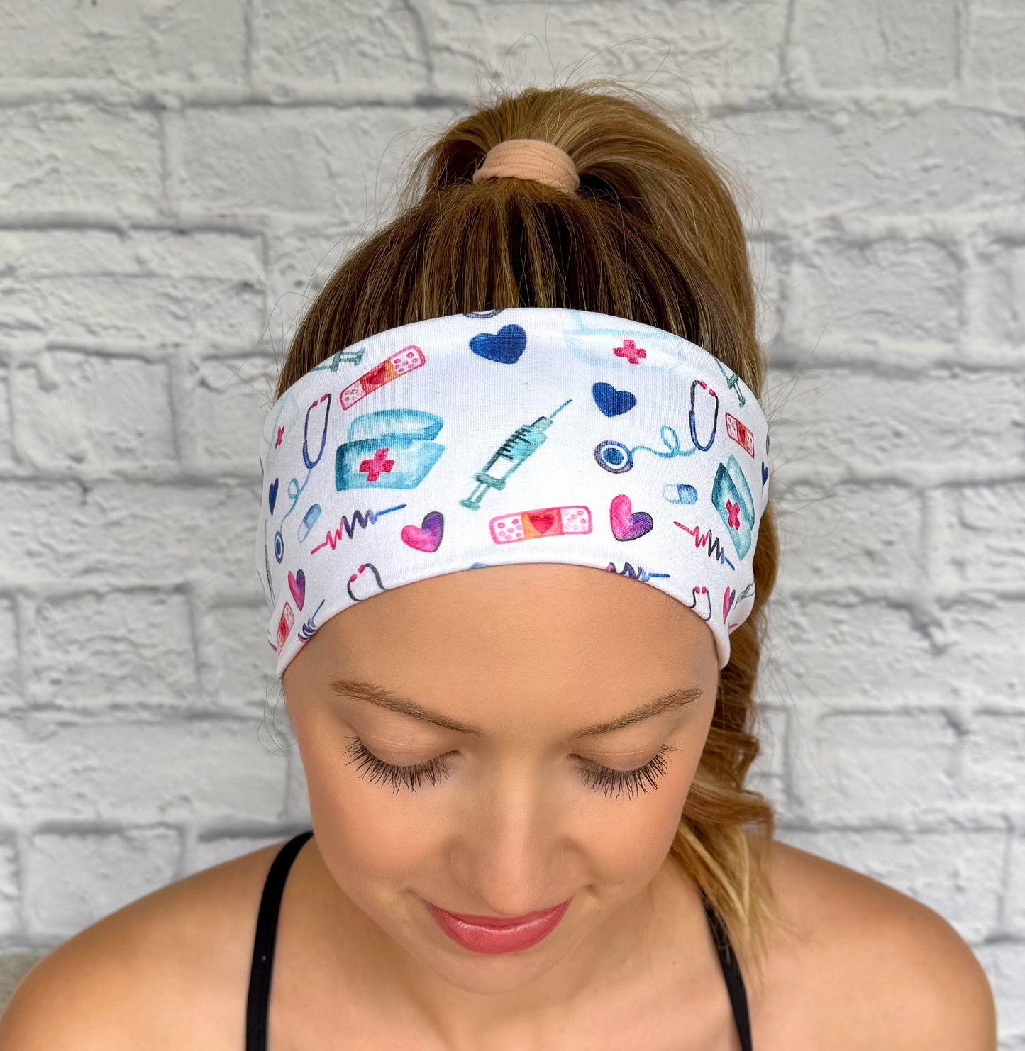 Woman with hair in ponytail wearing flat, stretchy, white headband with pink, blue, and purple medical theme print.