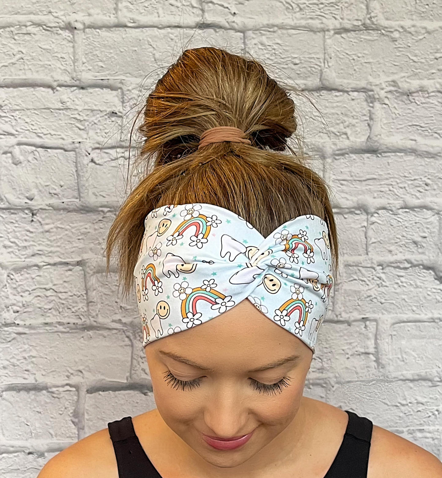 Woman with hair in bun wearing wide, twisted headband in light blue with teeth, rainbows, flowers, and smiley faces print.