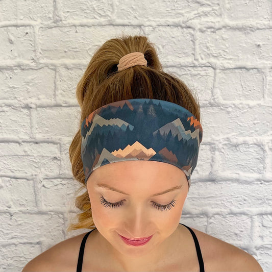 Woman with hair in curled ponytail wearing flat, stretchy headband in mountain print with varying shades of blue, brown, and gray.