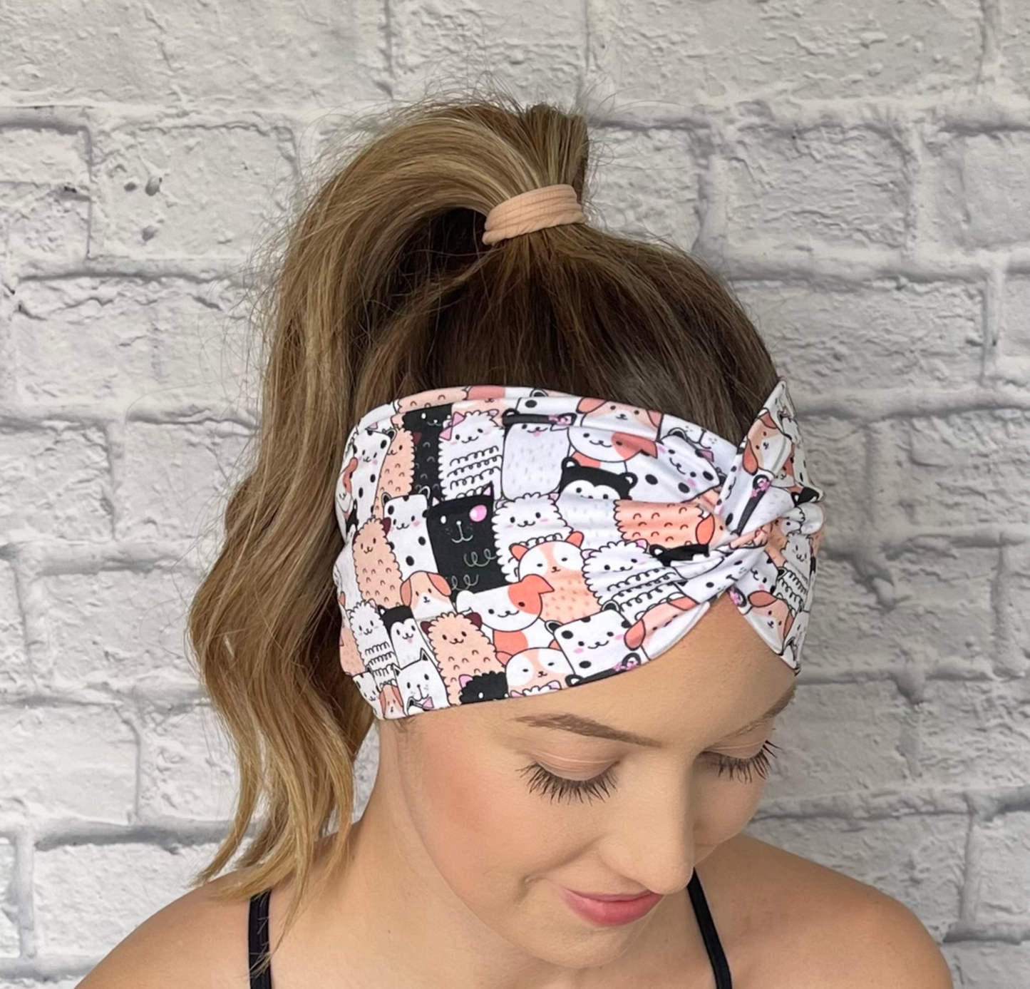 Woman with hair in curled ponytail wearing stretchy, wide, twisted headband with peach, white, and black animal print with dogs and cats.