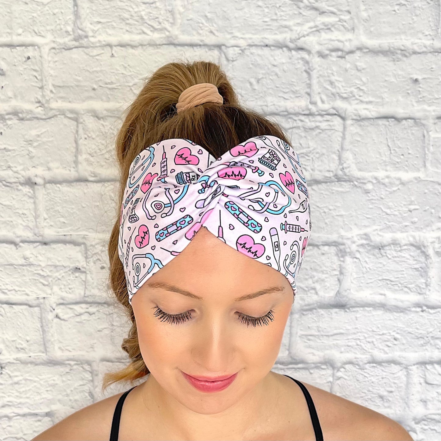 Woman with hair in curled ponytail wearing wide, twisted headband in light pink with pink, purple, and blue medical theme print.