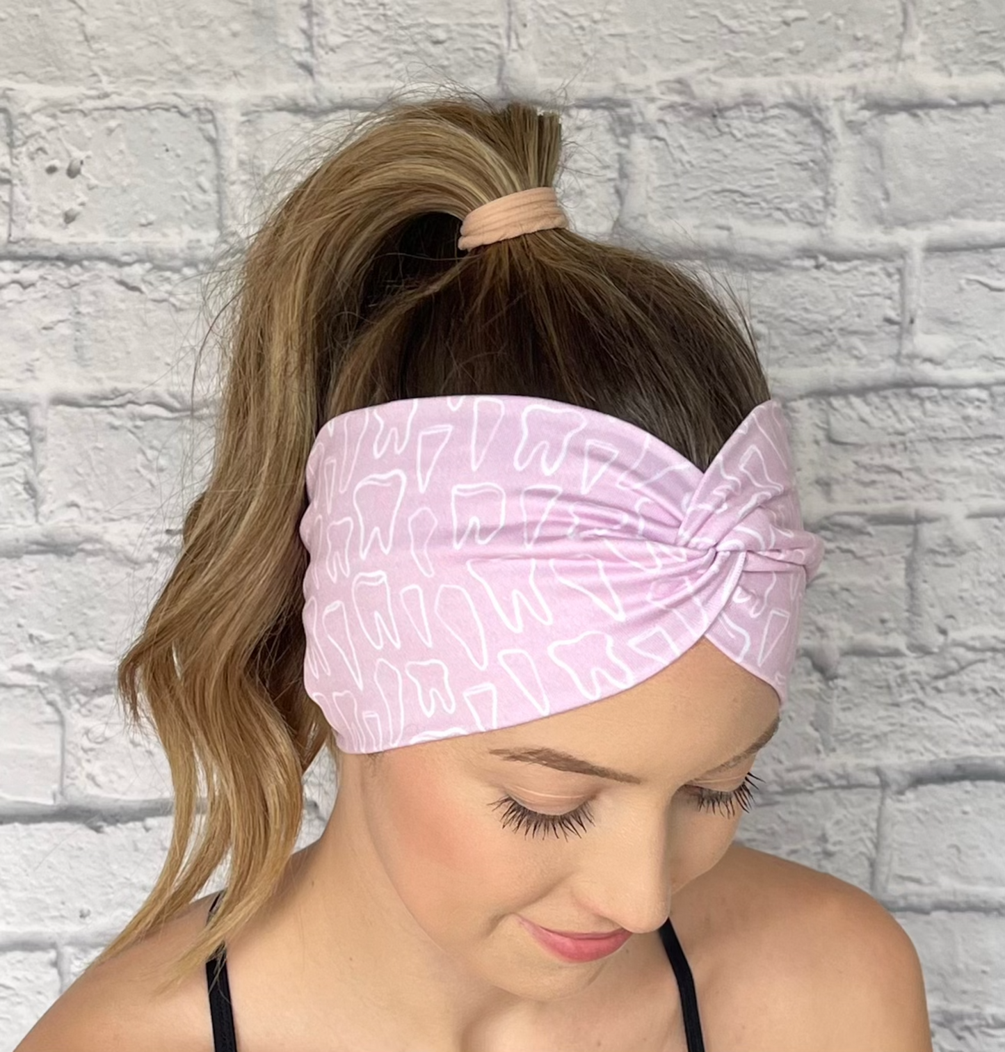 Woman with hair in curled ponytail wearing wide, twisted headband in light pink with white teeth print.