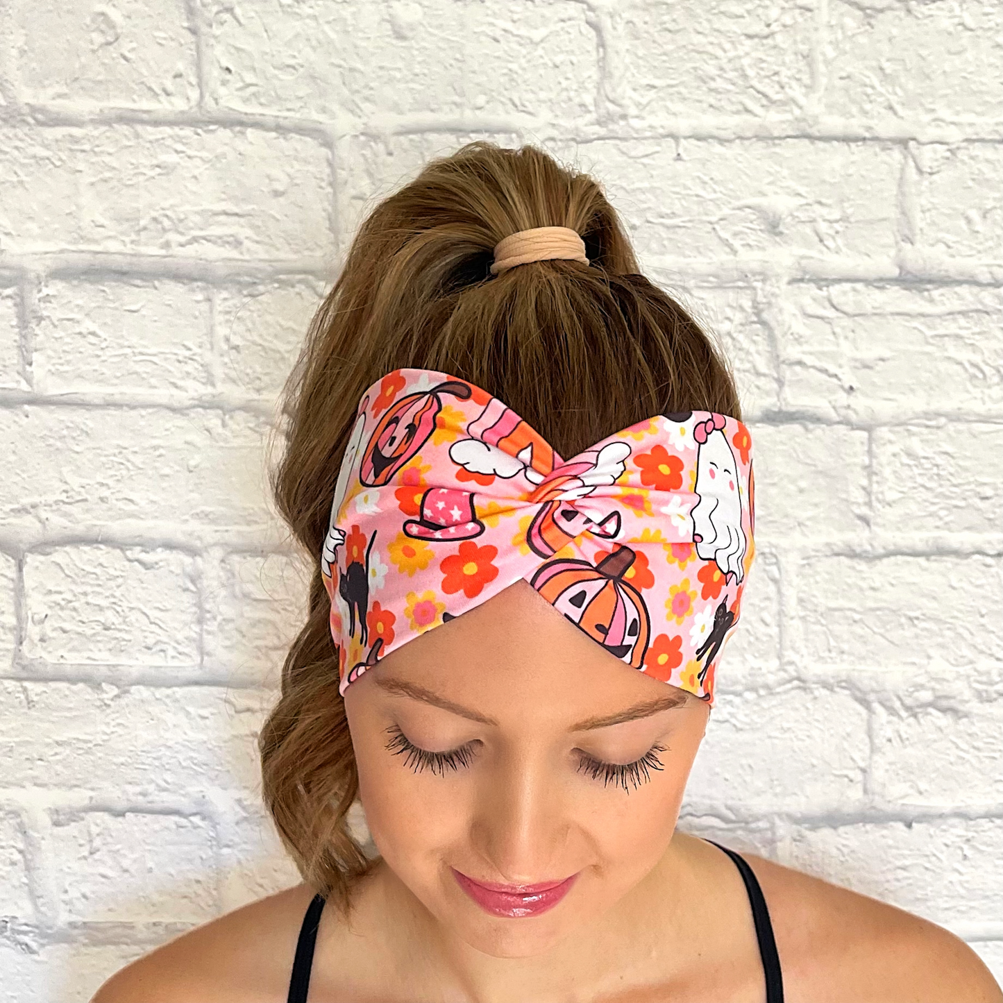 Woman with hair in curled ponytail wearing wide, twisted headband in pink with floral, rainbow, and halloween print in pink, white, black, yellow, and orange.