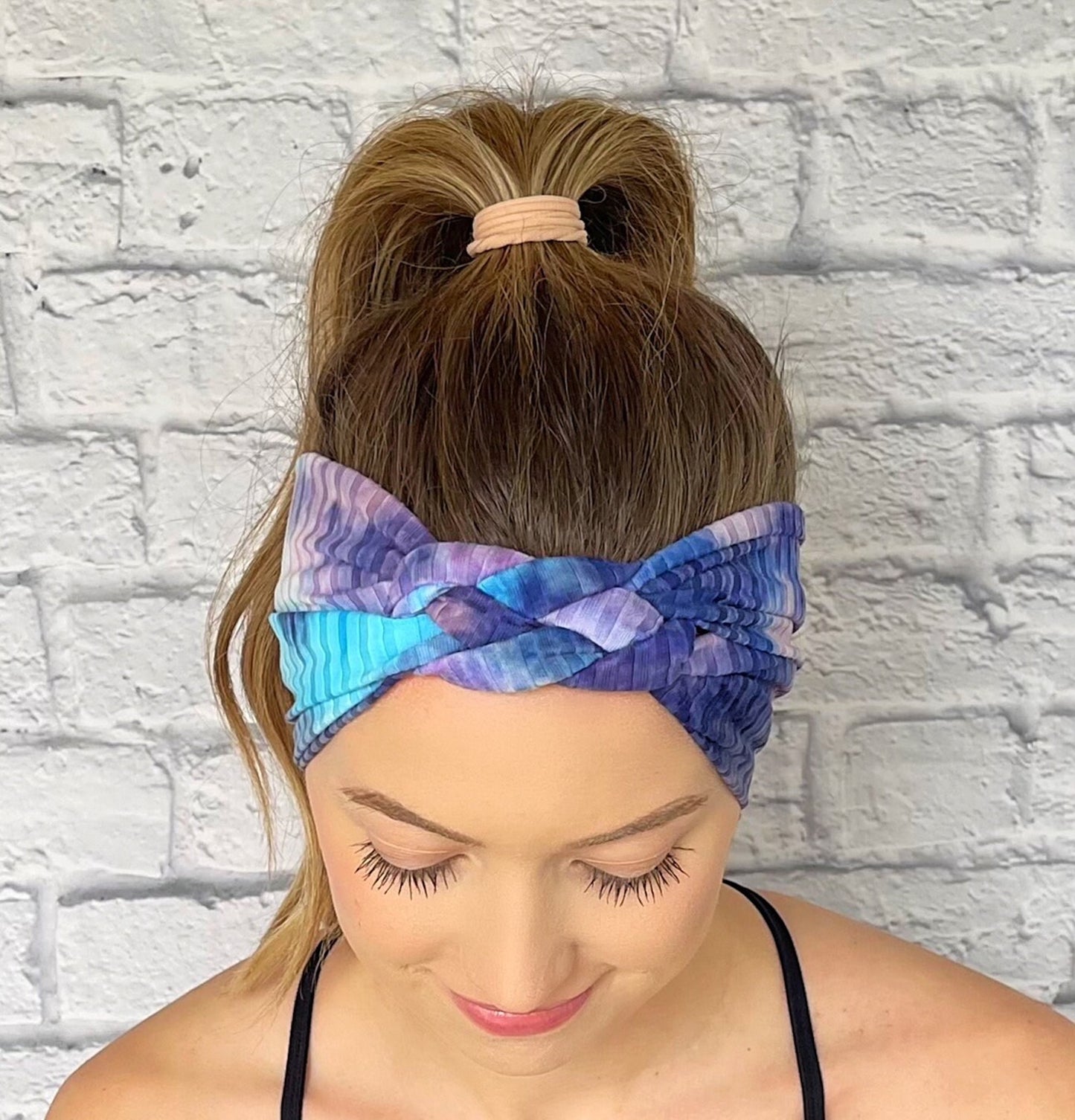 Woman with hair in curled ponytail wearing sailor knot style headband in ribbed fabric with purple and blue tie dye print.