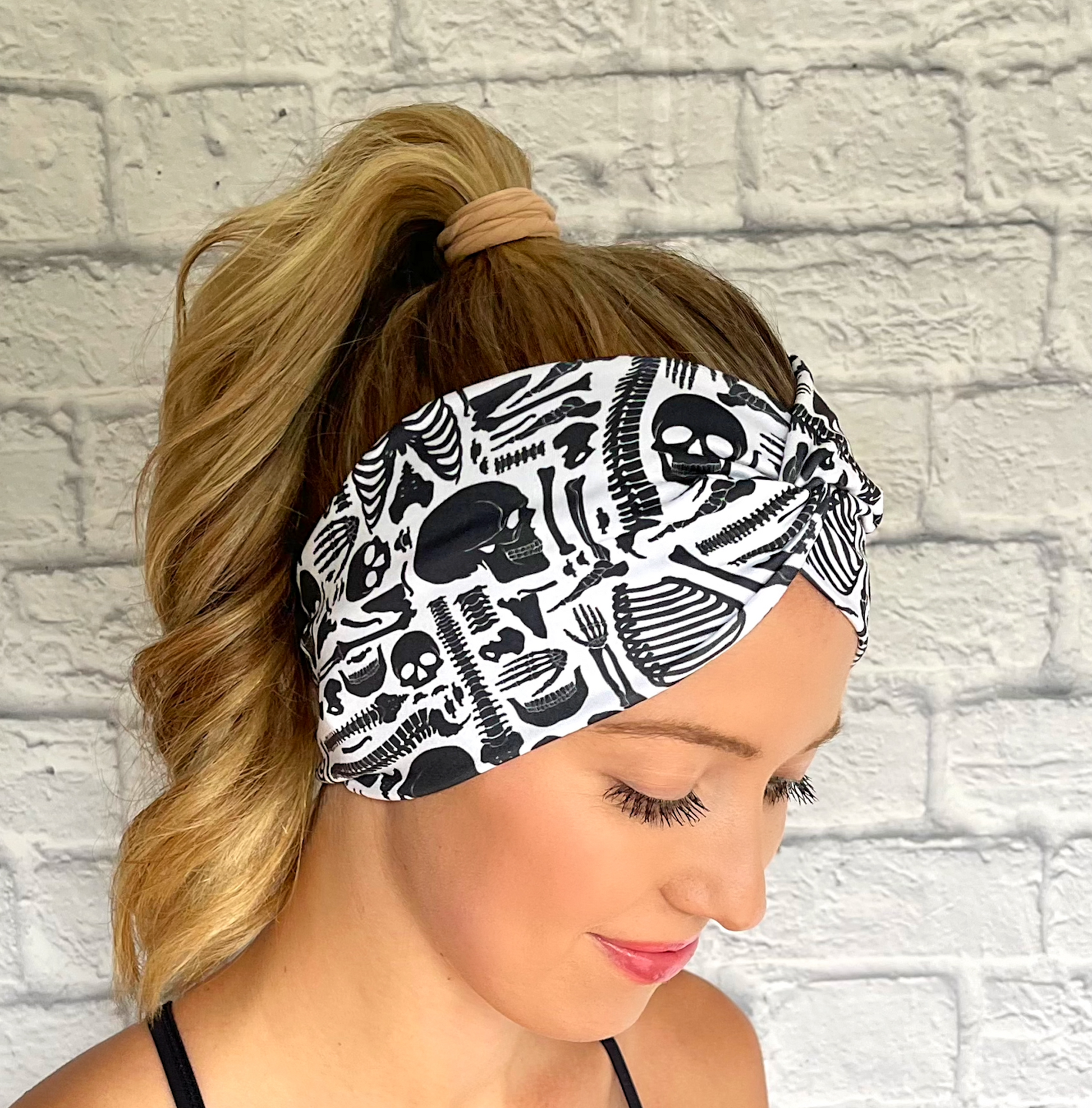 Woman with hair in curled ponytail wearing white, wide twist headband with black skeleton print.