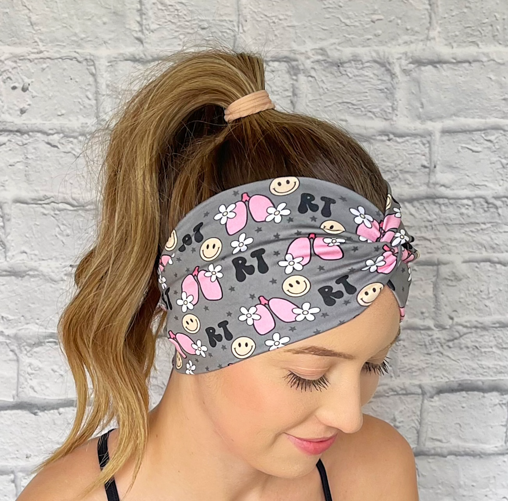 Woman with hair in curled ponytail wearing wide, twisted headband in gray with "RT" print, lungs, flowers, and smiley faces.