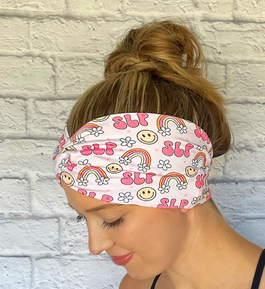 Woman with hair in bun wearing wide, twisted pink headband stating "slp", with rainbows, smiley faces, and white flowers.