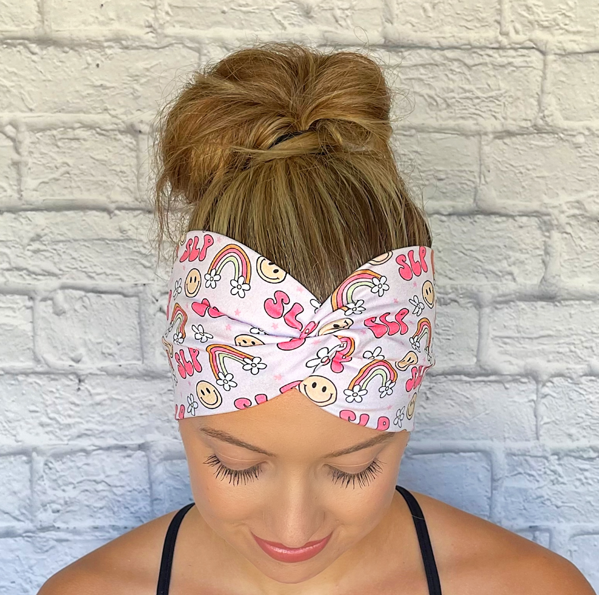Woman with hair in bun wearing wide, twisted pink headband stating "slp", with rainbows, smiley faces, and white flowers.