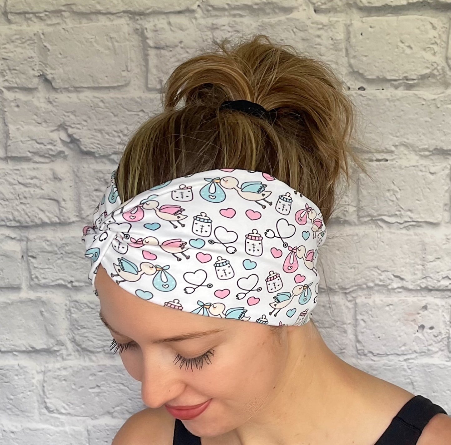 Woman with hair in bun wearing stretchy, wide, twisted headband in white with baby theme print- storks, hearts, bottles, and stethoscopes.