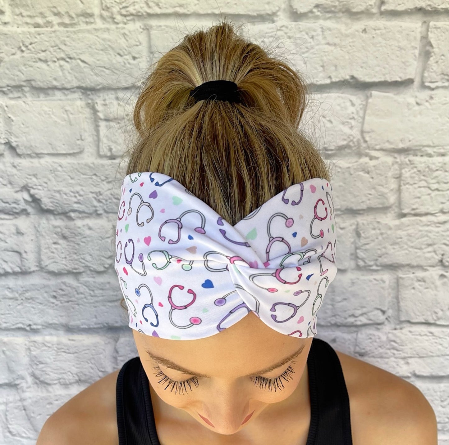 Woman with hair in ponytail wearing wide, twisted, white headband with pastel color stethoscope print.