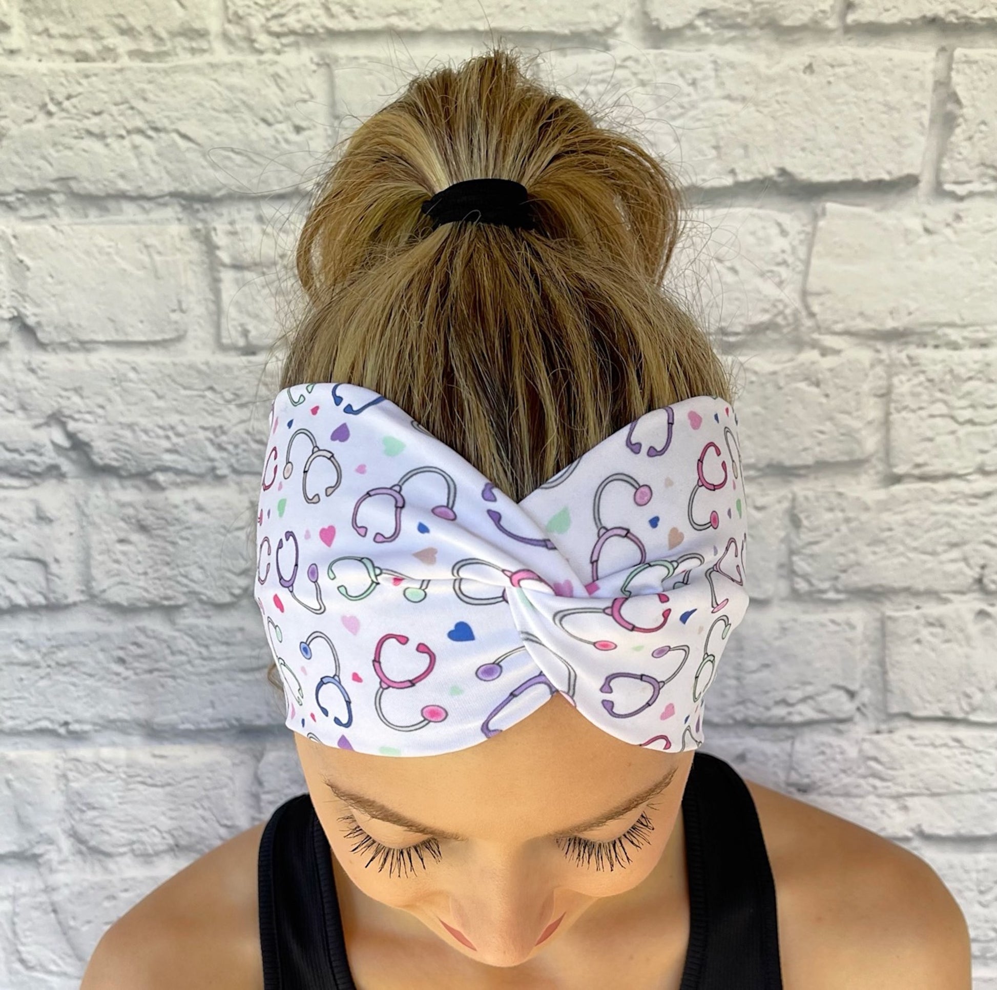 Woman with hair in ponytail wearing wide, twisted, white headband with pastel color stethoscope print.