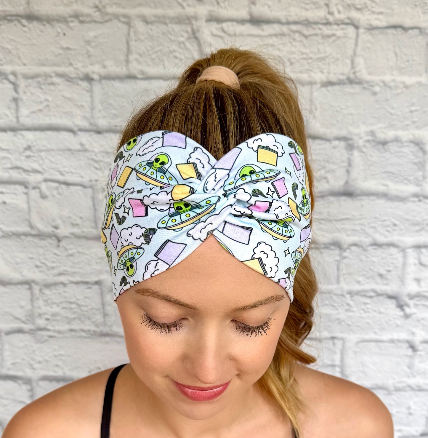 Woman with hair in ponytail wearing wide, twisted, light blue headband with alien theme print.