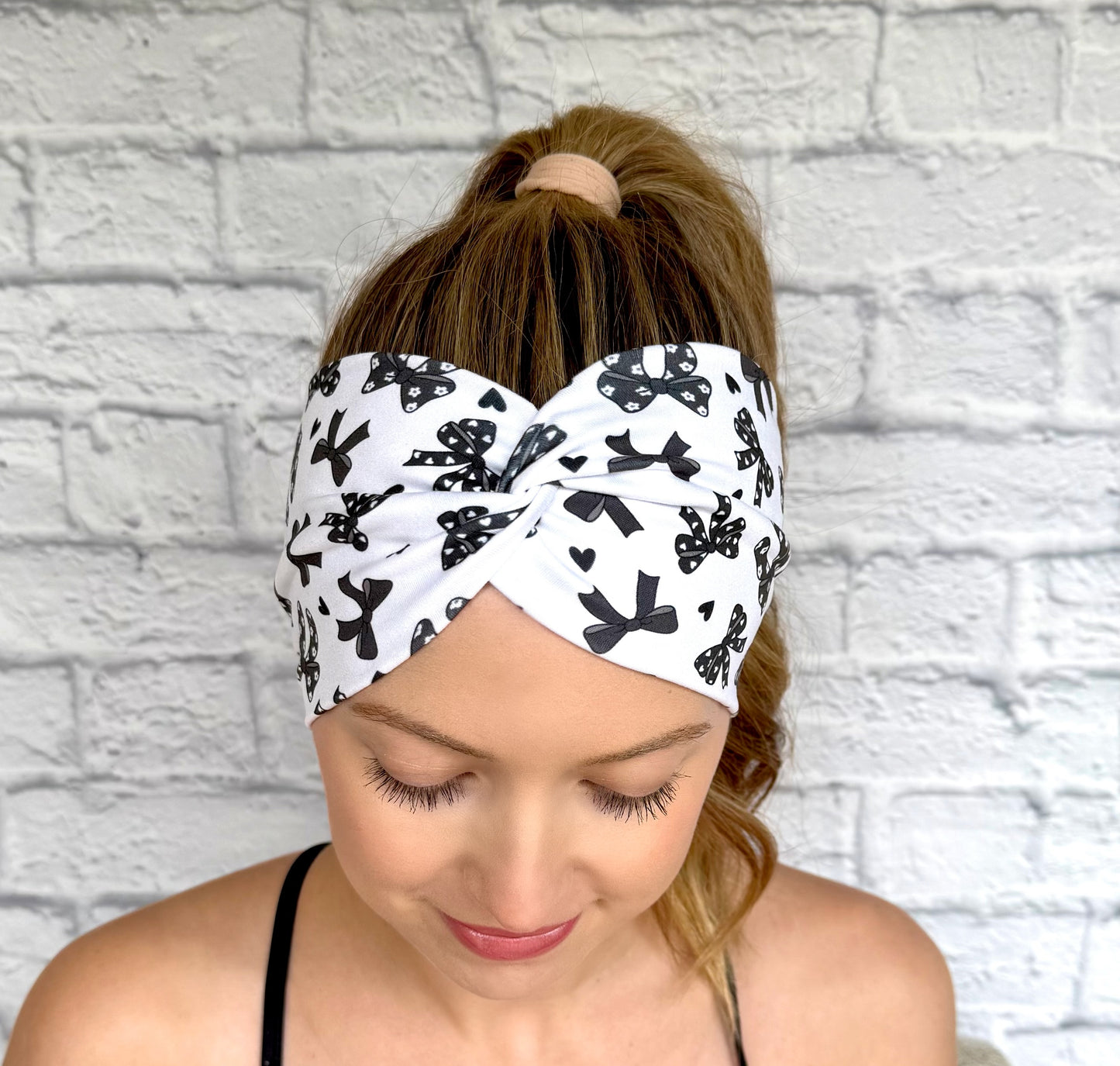 Woman with hair in curled ponytail wearing wide, twisted, white headband with black bow print.