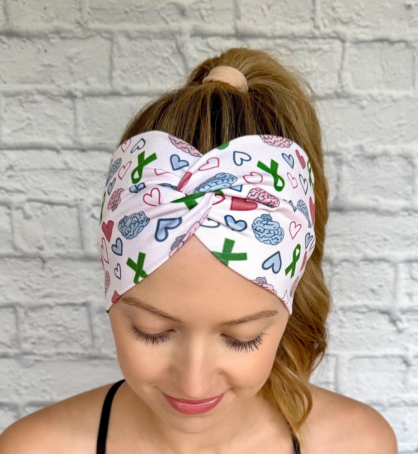 Woman with hair in curly ponytail wearing wide, twisted headband with green ribbon, pink and blue brain, and pink and blue heart print.