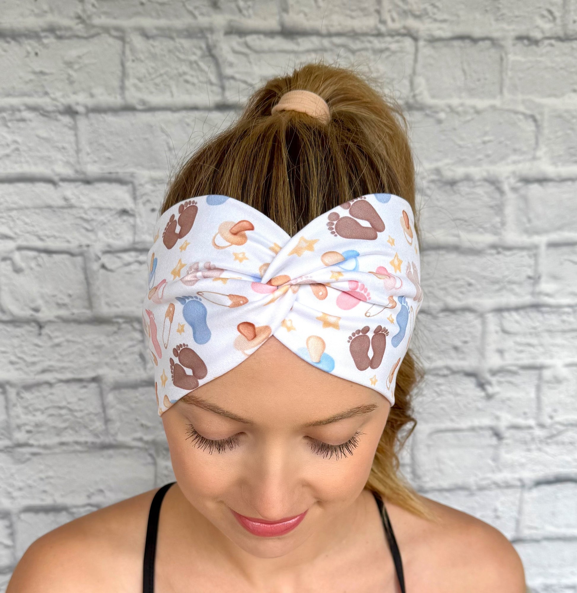 Woman with hair in ponytail wearing wide, twisted, white headband with baby feet and baby theme print.
