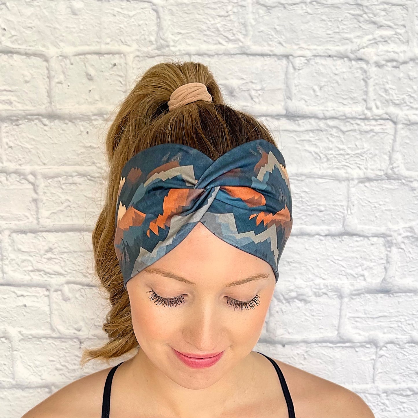 Woman with hair in curled ponytail wearing wide, twisted headband with mountain print in varying shades of blue, brown, and gray.