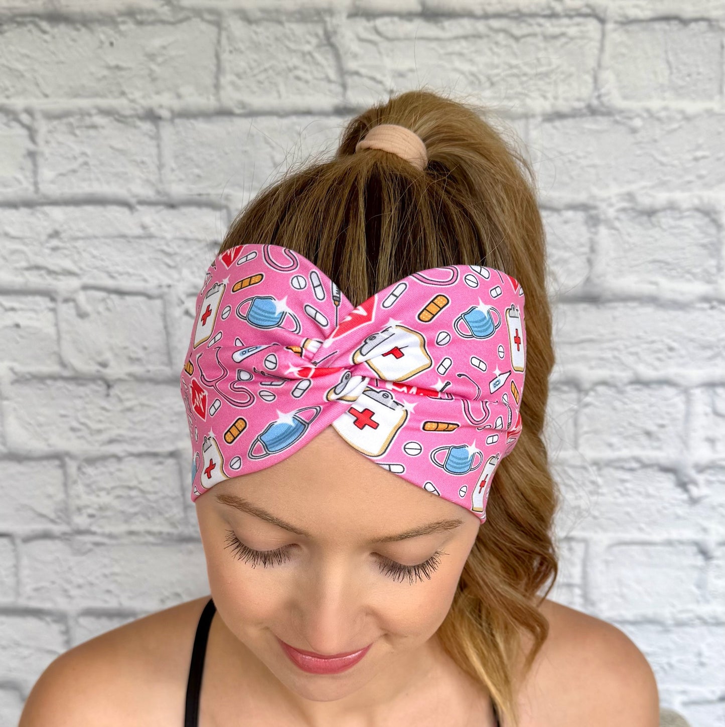 Woman with hair in ponytail wearing wide, twisted, stretchy headband in pink with medical theme print.
