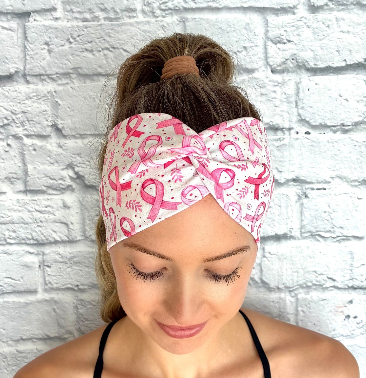 Woman with hair in ponytail wearing wide, twisted, white/cream headband with pink ribbon and pink floral print against a white brick wall.