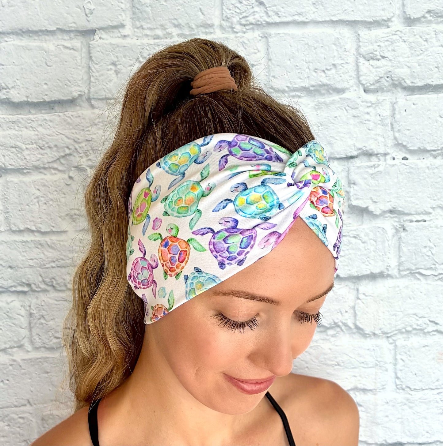 Woman with hair in ponytail wearing wide, twisted, white headband with sea turtle print, displayed on white brick background.
