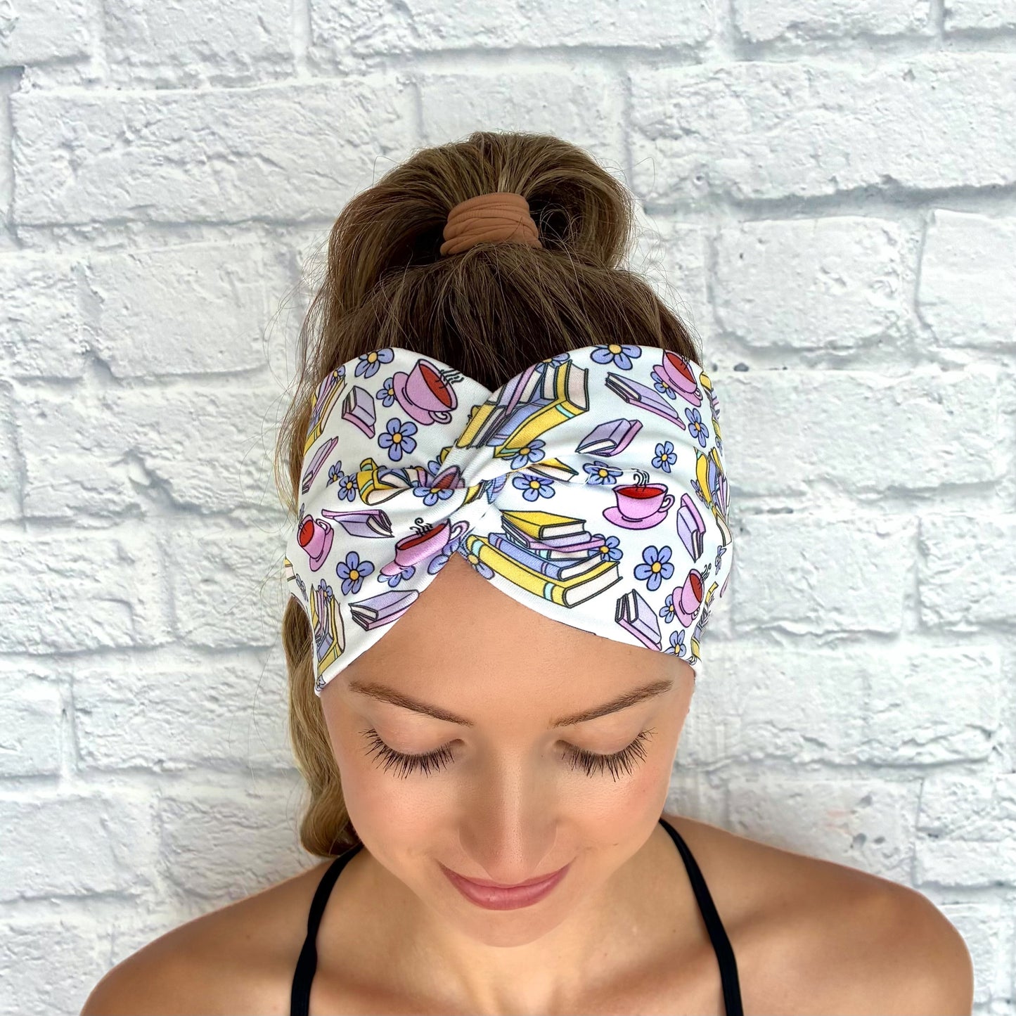 Woman with hair in ponytail wearing wide, twisted, white headband with coffee, flower, and book print.