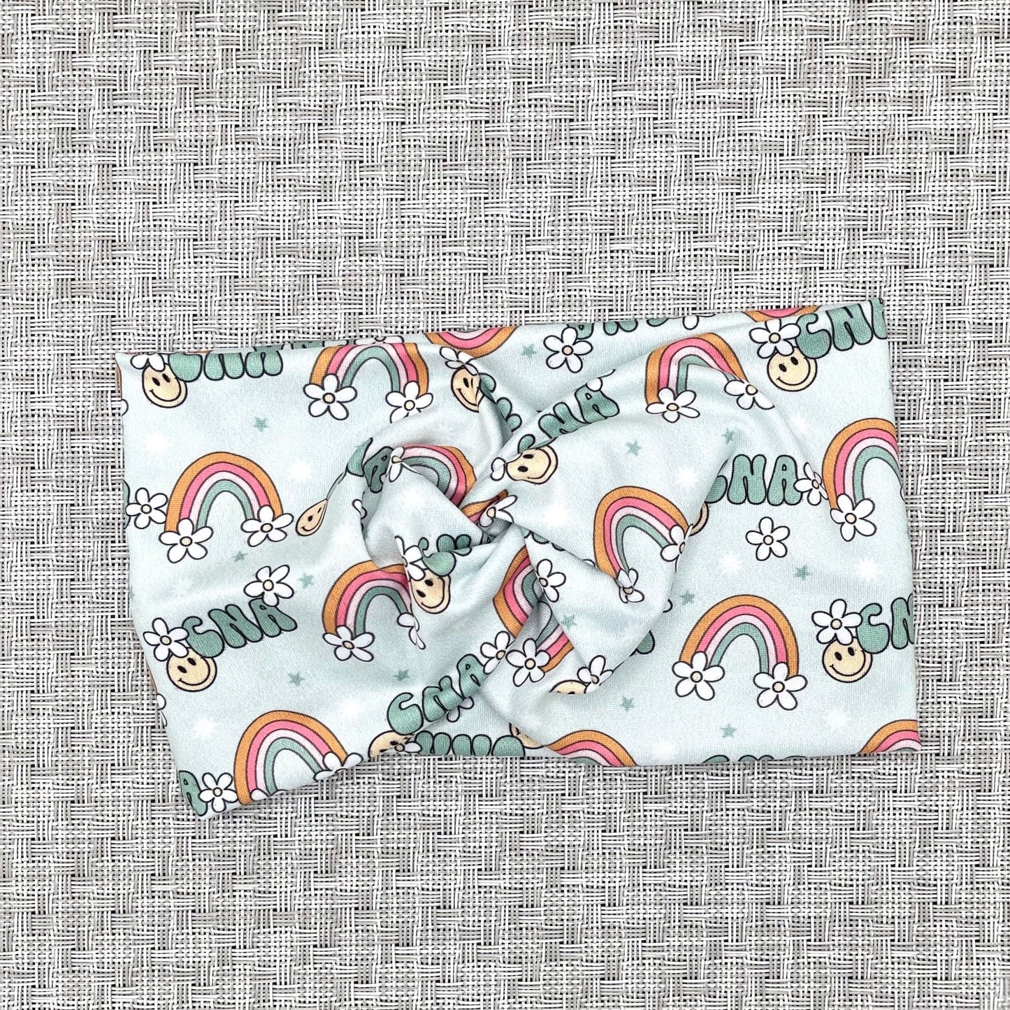 Wide, twisted, light blue headband with "cna" print, rainbows, white flowers, and smiley faces.
