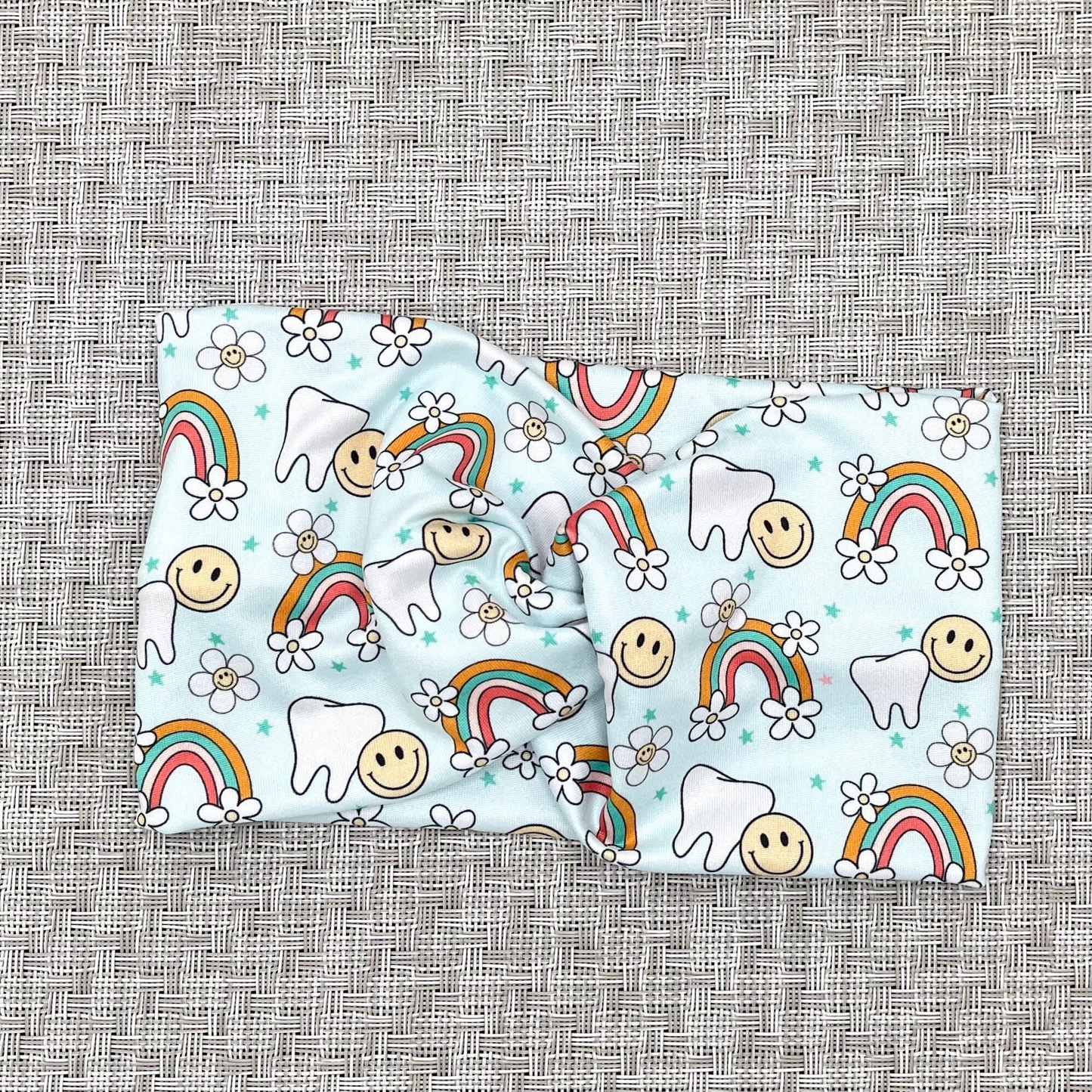 Wide, twisted headband in light blue with teeth, rainbows, flowers, and smiley faces print.
