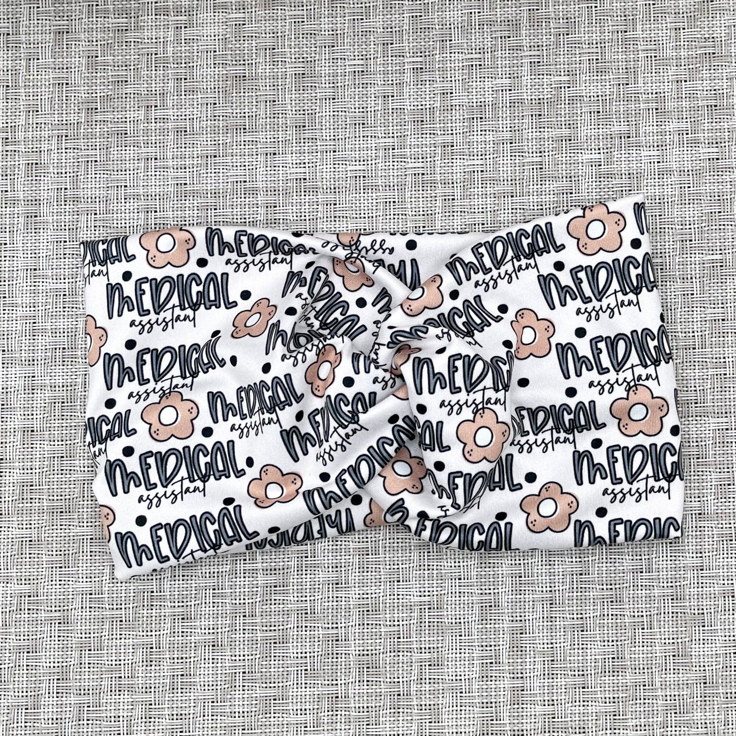 Cream, wide, twisted headband with "medical assistant", flowers, and polka dot print in blue and tan.