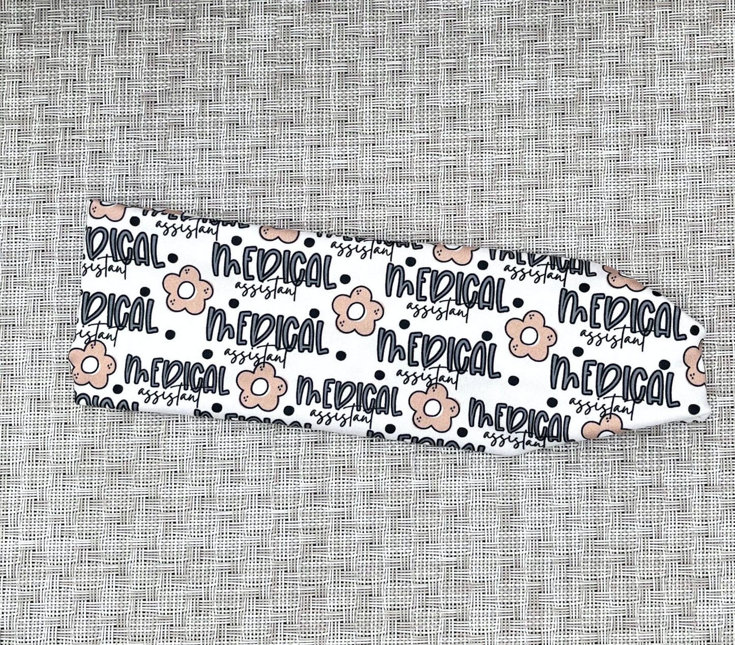 Cream, stretchy headband with "medical assistant" print in navy blue, black polka dots, and tan flowers.