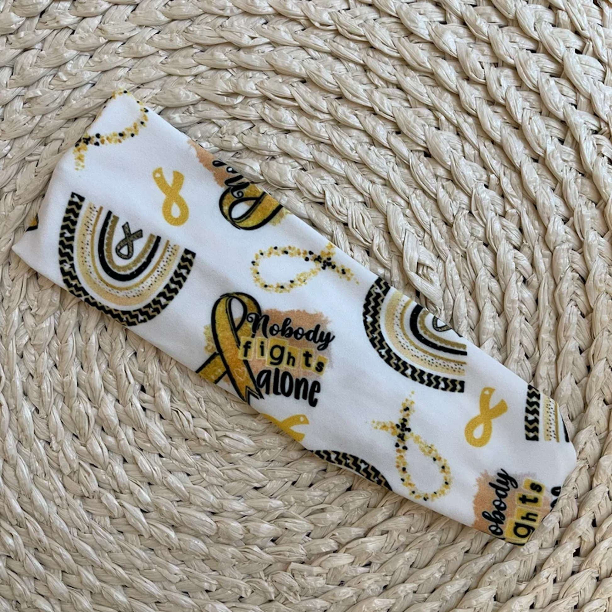 Flat, stretchy headband in white with "no one fights alone" print, yellow/gold rainbow, and yellow/gold ribbon print.