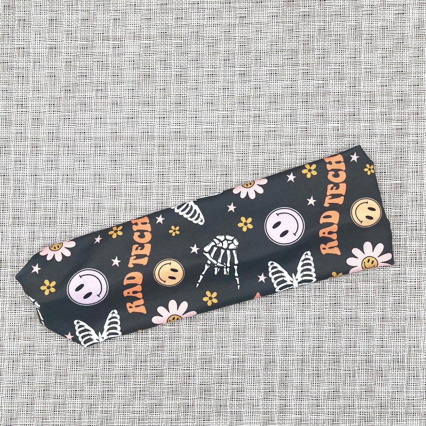 Flat, stretchy, gray headband with "rad tech", smiley faces, flowers, and skeletons print.