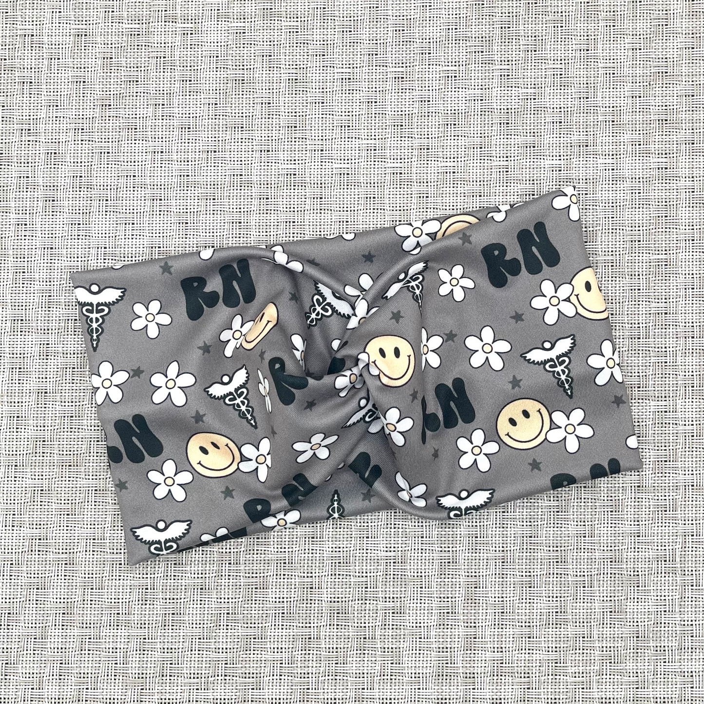Wide, twisted headband. The headband is gray with "RN" print, flowers, smiley faces, and medical theme print in black, white, and yellow.